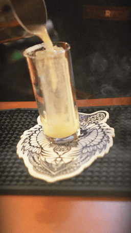 A tall glass with a cocktail, ice, and a lemon wedge, with a straw inside, placed on a decorative napkin and black tray.