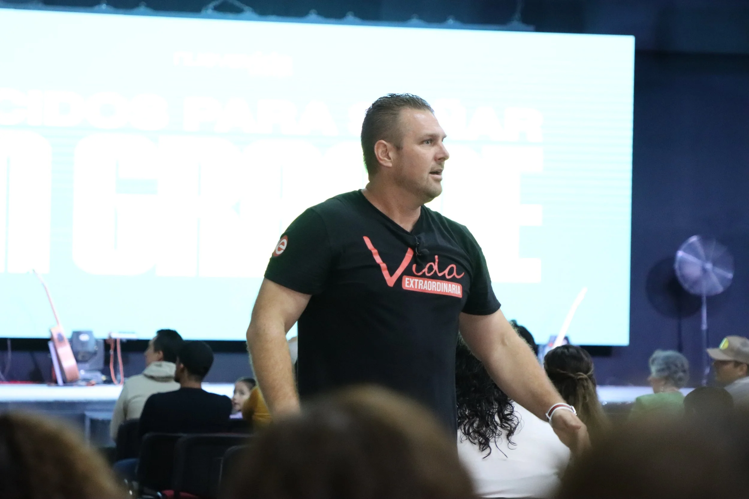Un hombre con camiseta negra con letras rojas y blancas, sentado en un auditorio con varias personas, en un escenario con una pantalla grande de fondo.