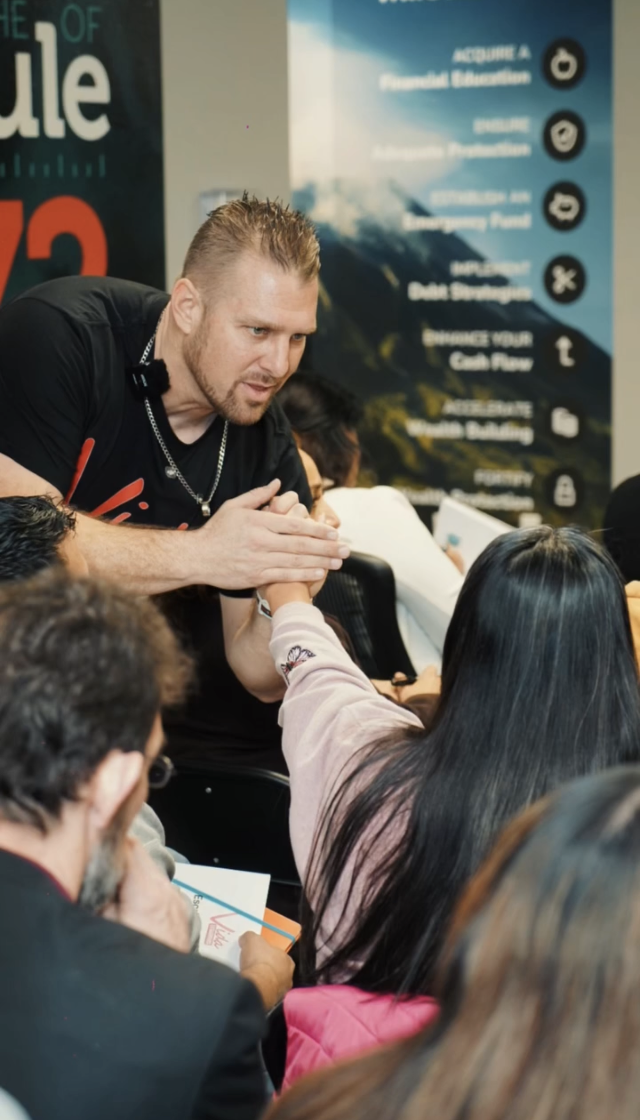 Persona interactuando con un grupo de personas en una conferencia o taller, en un entorno interior con cartelera de fondo que muestra esquemas y frases relacionadas con educación y financiamiento.