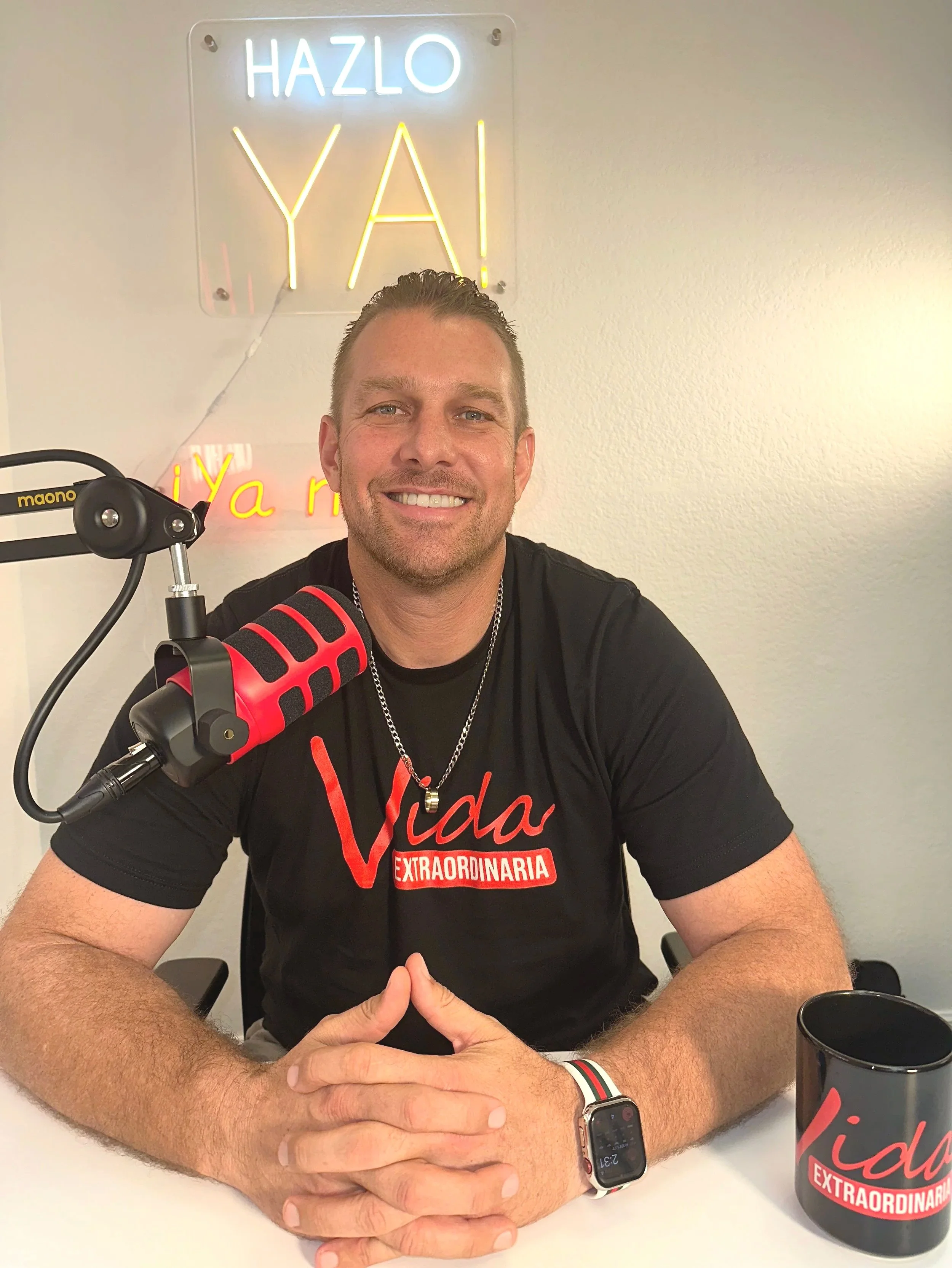Hombre sonriendo en un estudio, con micrófono rojo y negro y taza con la palabra 'Vida Extraordinaria', en una pared con cartel de neón que dice 'Hazlo YAI'.