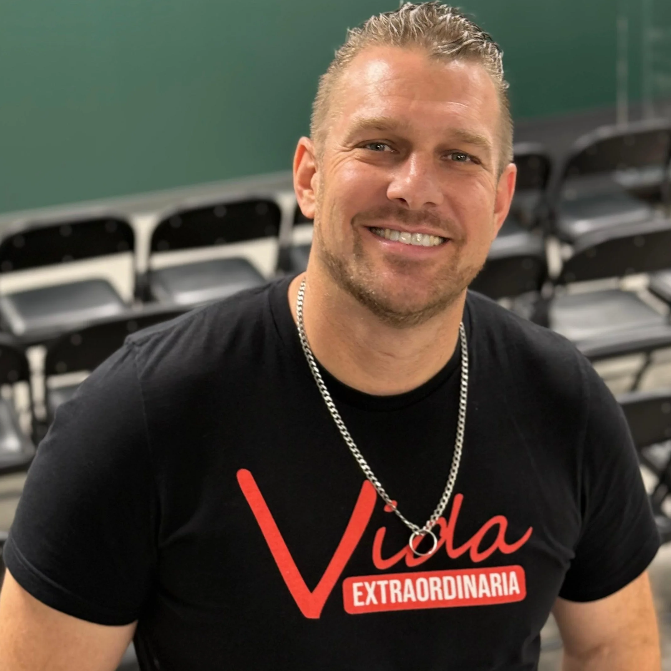 Un hombre sonriendo, con cabello rubio y peinado hacia atrás, viste una camiseta negra con texto rojo que dice 'Viva Extraordinaria', y lleva una cadena plateada. Está en un lugar con sillas negras apiladas en el fondo.