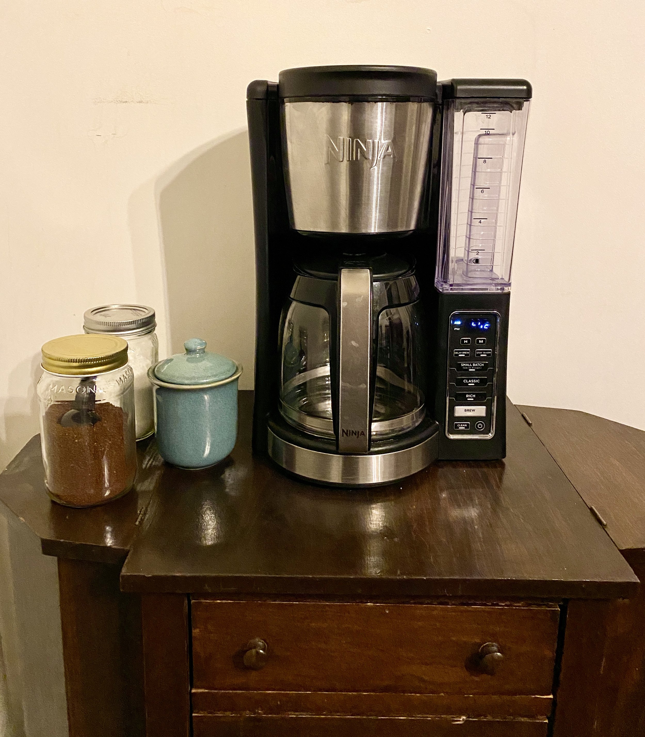 A coffee maker on a dark wooden table with jars and a small ceramic container beside it.