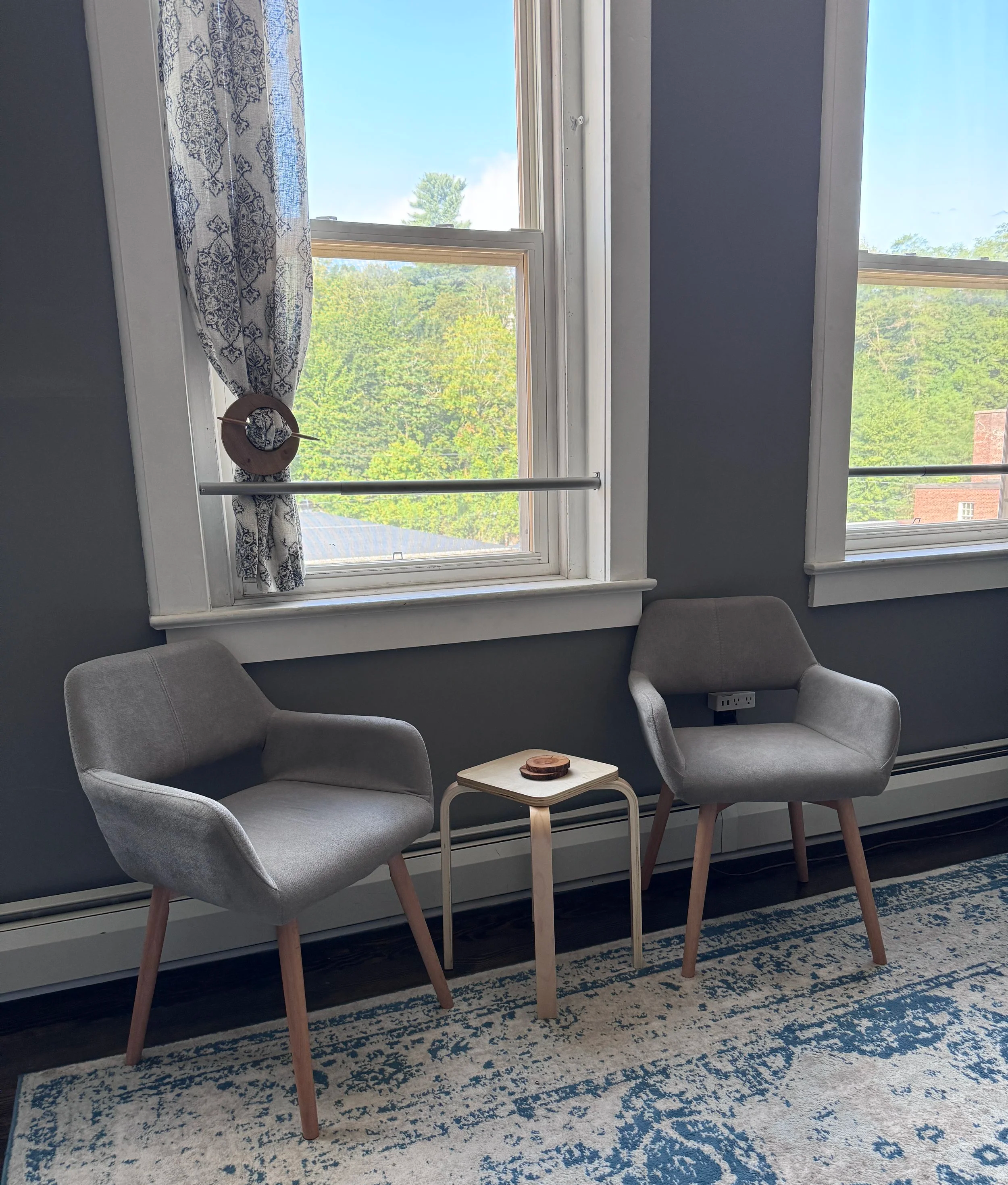 Two gray armchairs with wooden legs and a small white side table between them, set against a blue wall with three large windows showing green trees outside, and decorated with a patterned curtain and a circular decoration on the window.