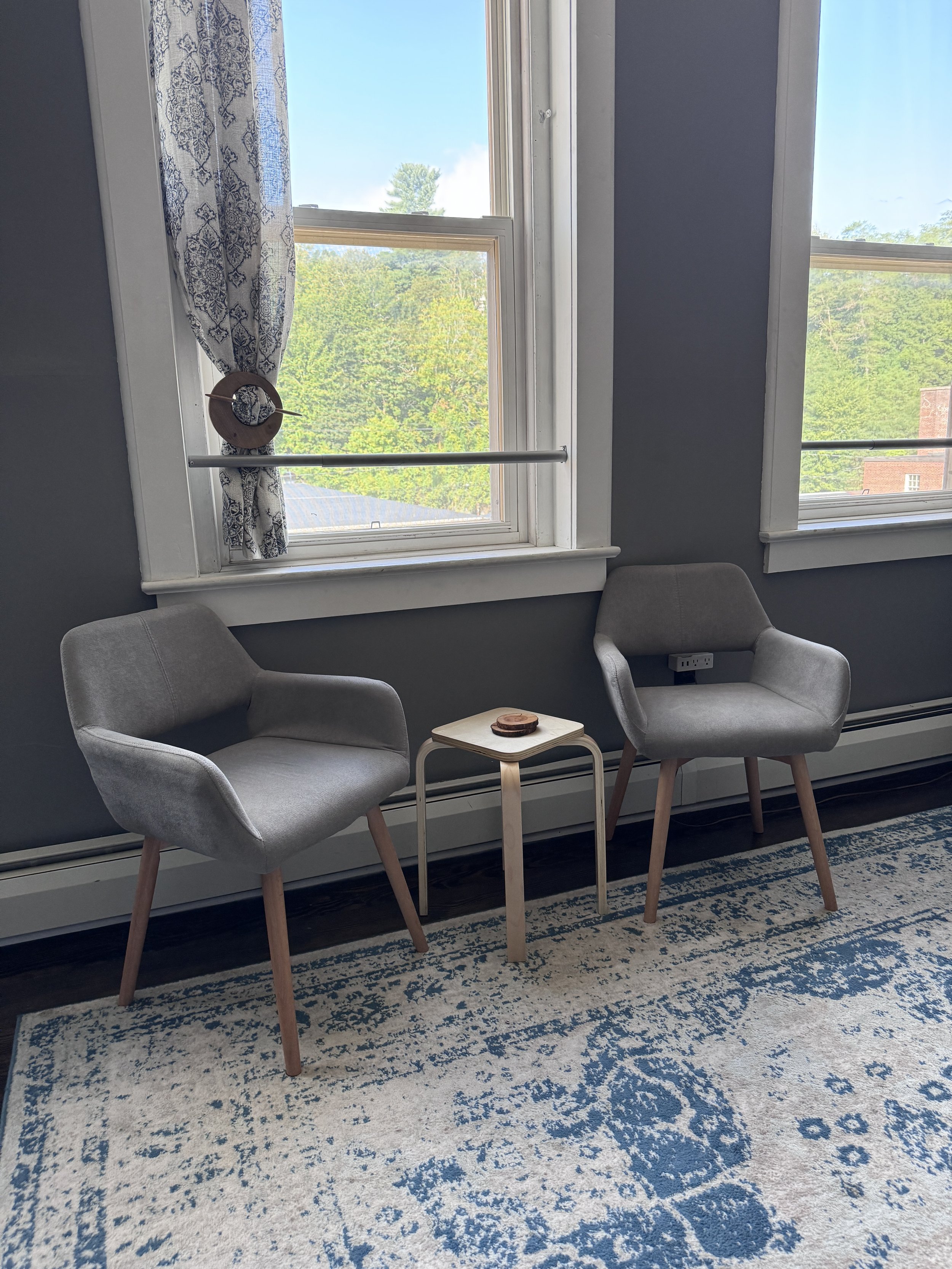 Two gray armchairs with wooden legs and a small white side table between them, set against a blue wall with three large windows showing green trees outside, and decorated with a patterned curtain and a circular decoration on the window.