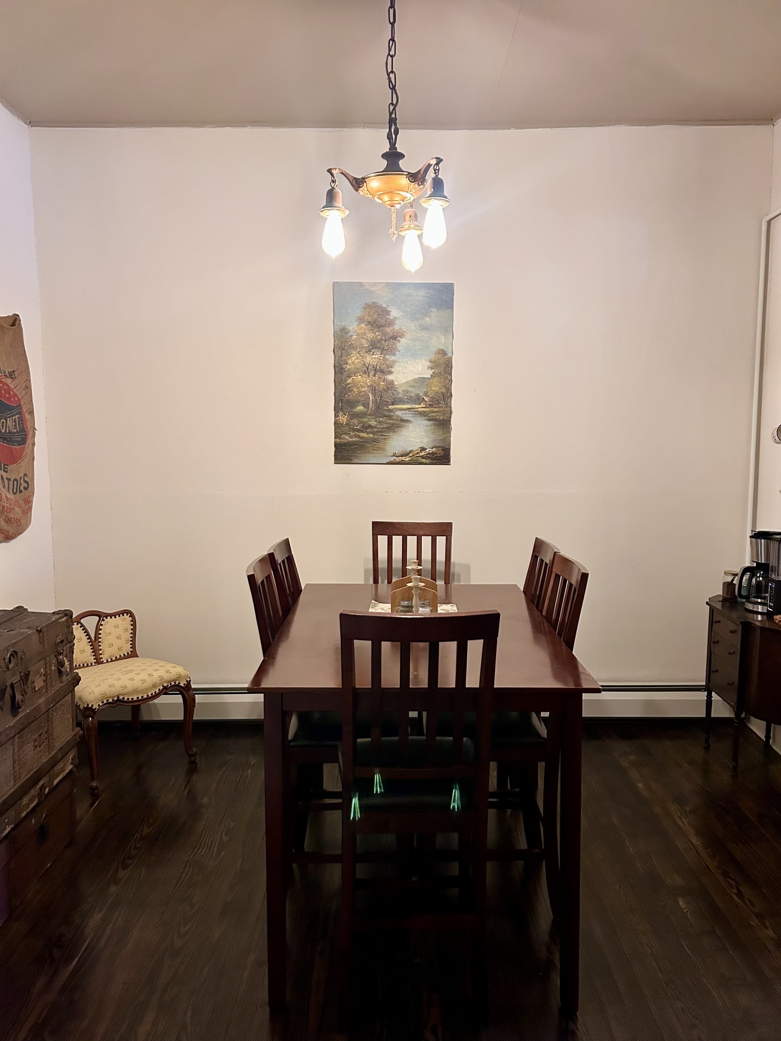 A dining room with a wooden table and six matching wooden chairs, a chandelier hanging from the ceiling, a landscape painting on the wall, and a sideboard with a coffee maker and other small items.