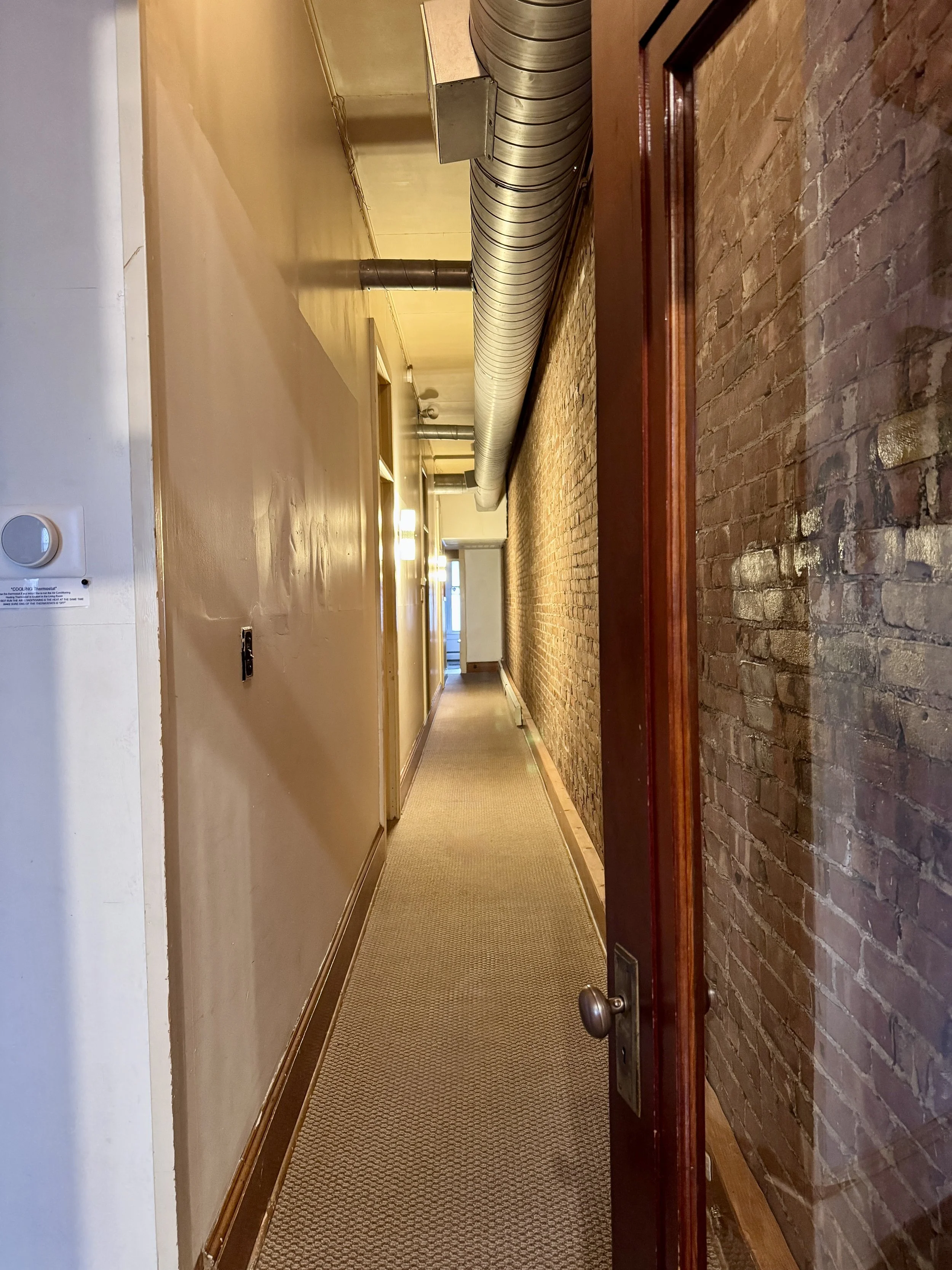 A view of a long, narrow hallway with beige and brick walls, carpeted floor, and an exposed industrial ventilation duct running along the ceiling. The hallway is illuminated by wall-mounted lights and extends towards a door at the end.