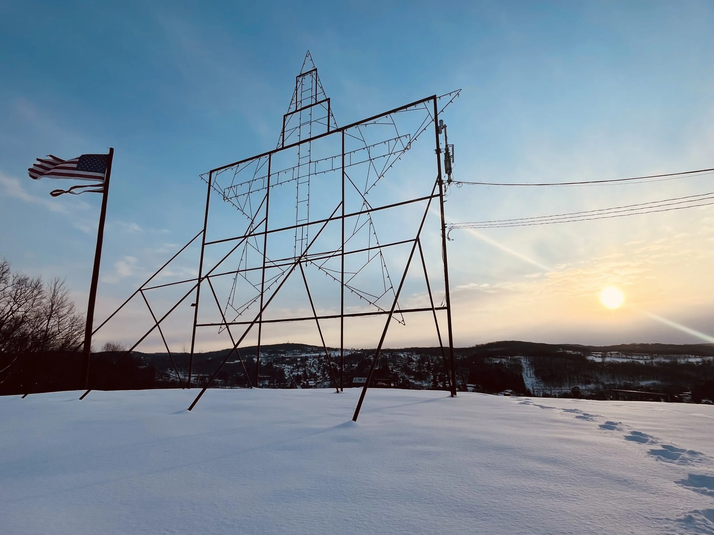 Snow-covered landscape with a large star-shaped metal frame and an American flag waving in the wind at sunset.