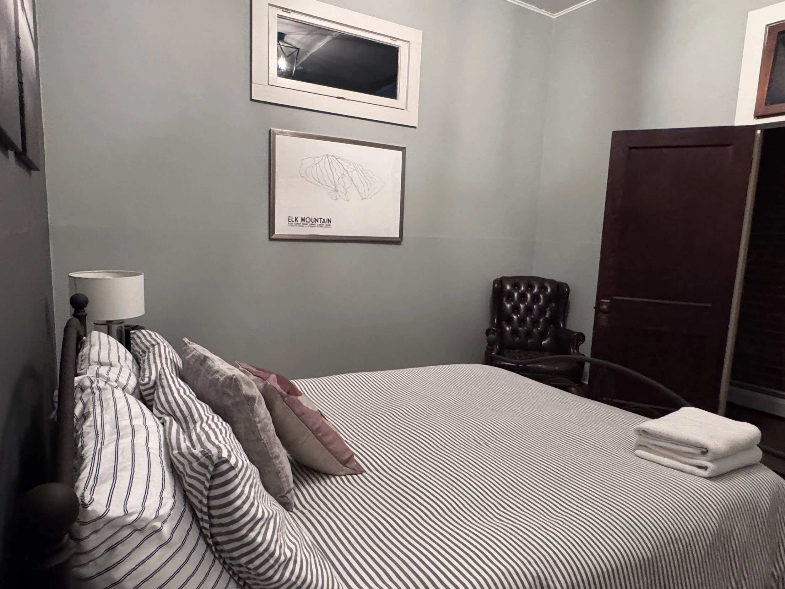 A cozy bedroom featuring a bed with striped bedding and several pillows, a bedside lamp, a framed mountain map poster, a window, a dark wooden wardrobe, and a leather armchair in the corner.