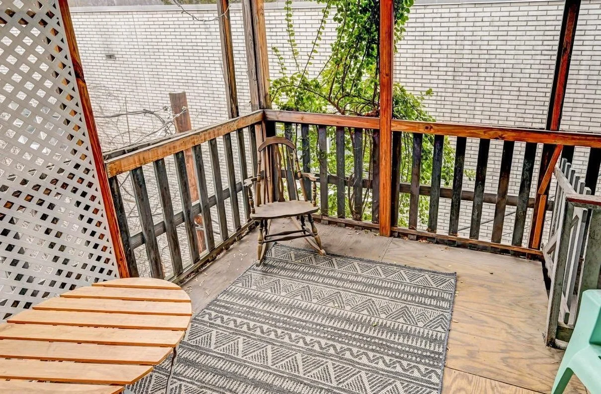 A small outdoor balcony with a wooden railing, two chairs, a round wooden table, a patterned outdoor rug, a plant climbing the railing, and a brick wall in the background.