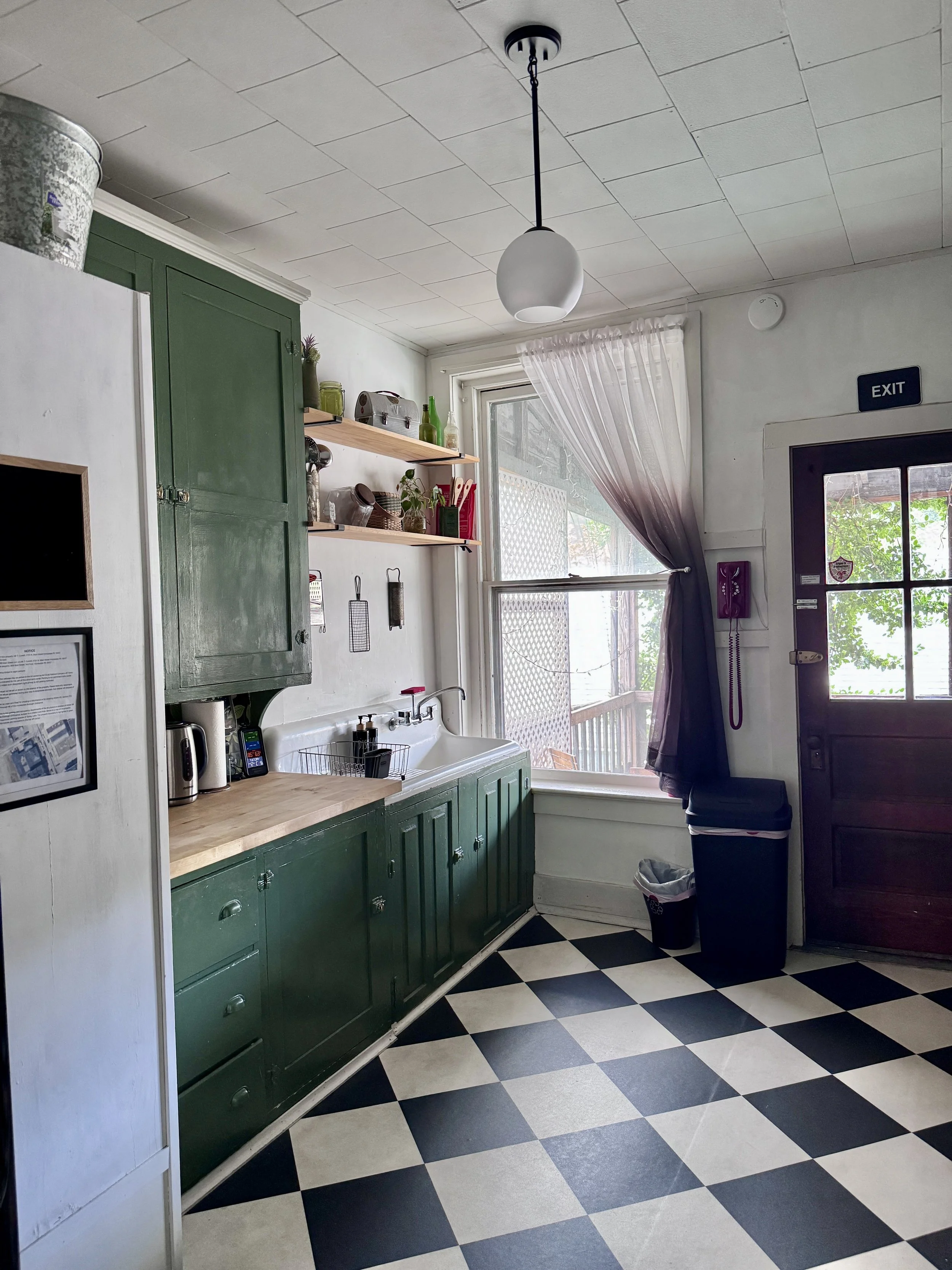 Kitchen with green cabinets, black and white checkered floor, window with curtain, and a door with an exit sign. Items on the shelves include bottles, potted plants, kitchen utensils, and a small microwave. There is a white sink under the window and a trash can near the door.