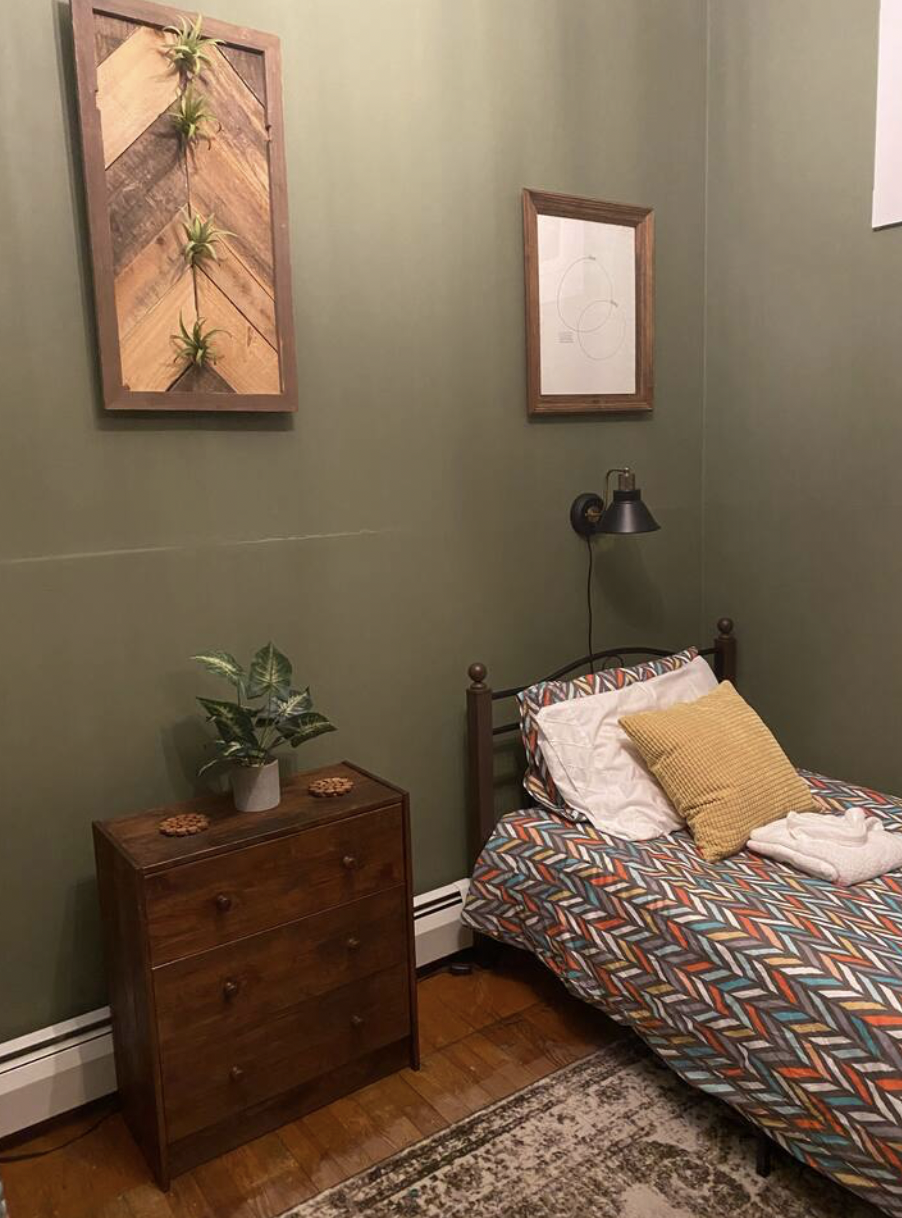 Bedroom with green walls, a wooden dresser with a plant, framed artwork, a bed with colorful geometric bedding, pillows, and a towel, and a wall-mounted lamp.