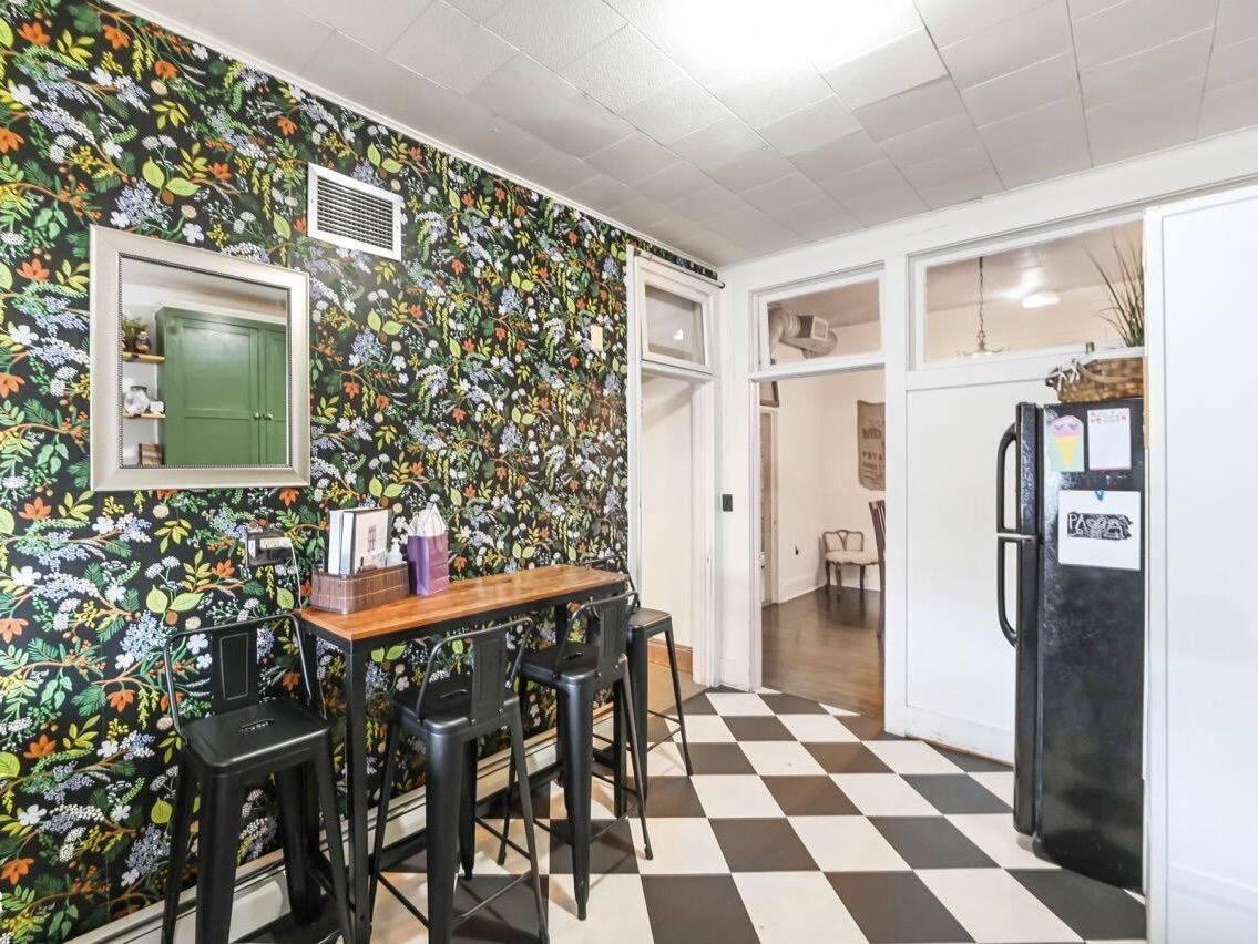 A kitchen corner with a black and white checkered floor, a floral wallpapered wall, a small wooden table with three black stools, a mirror on the wall, and a black refrigerator.