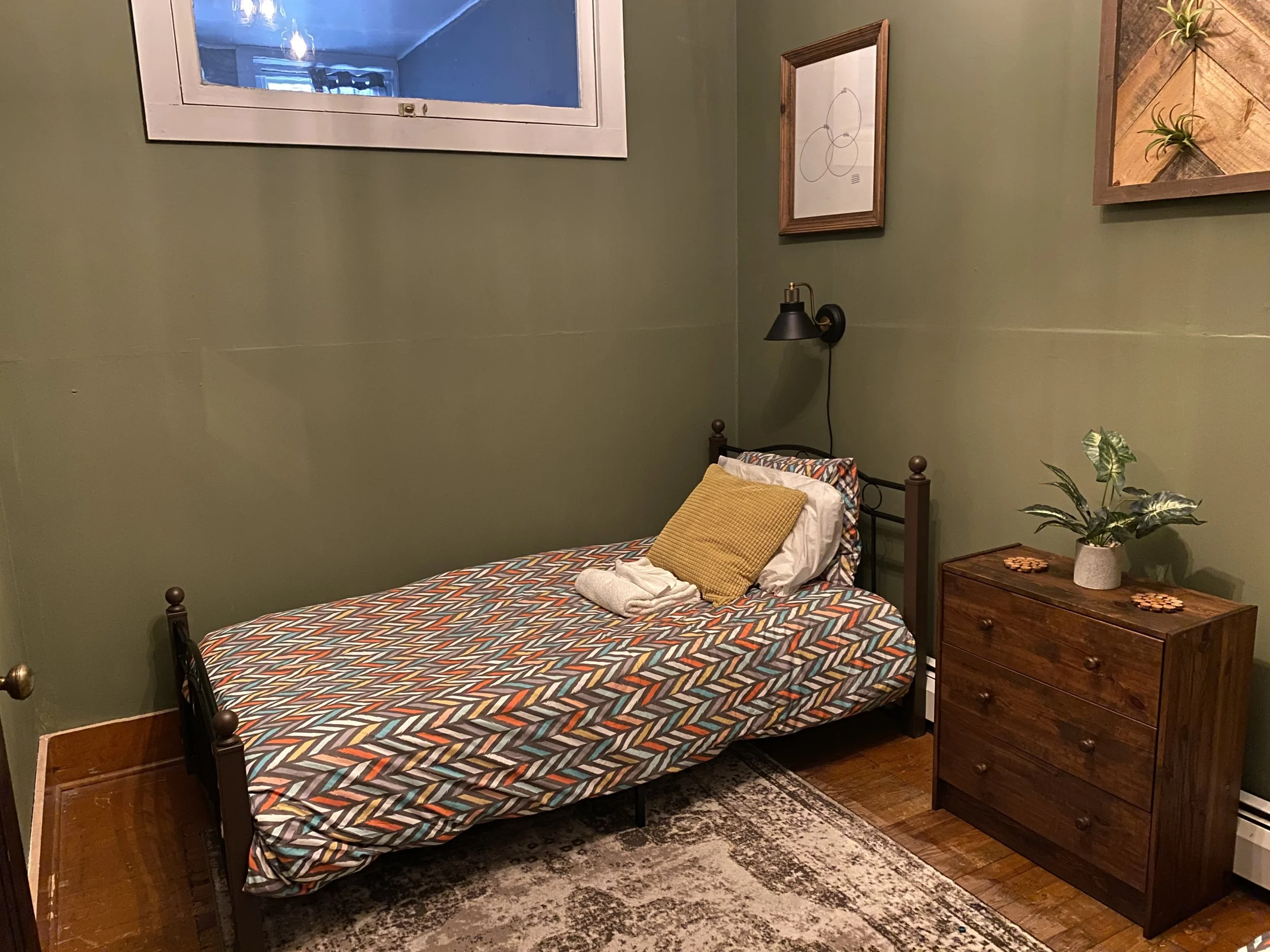 A small bedroom with a single bed, a wooden dresser, a potted plant, and framed artwork on sage green walls. The bed has a colorful zigzag-patterned bedsheet, a yellow pillow, and some folded clothes. A black wall-mounted lamp is above the bed.