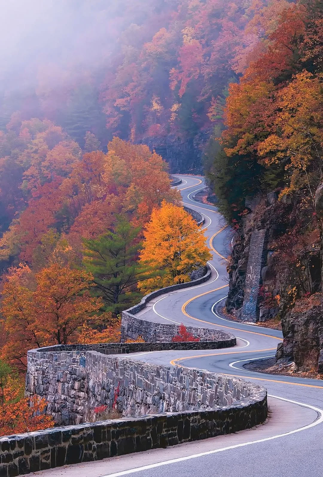 A winding mountain road with a stone wall on one side, surrounded by colorful fall foliage trees and a foggy hillside in the background.