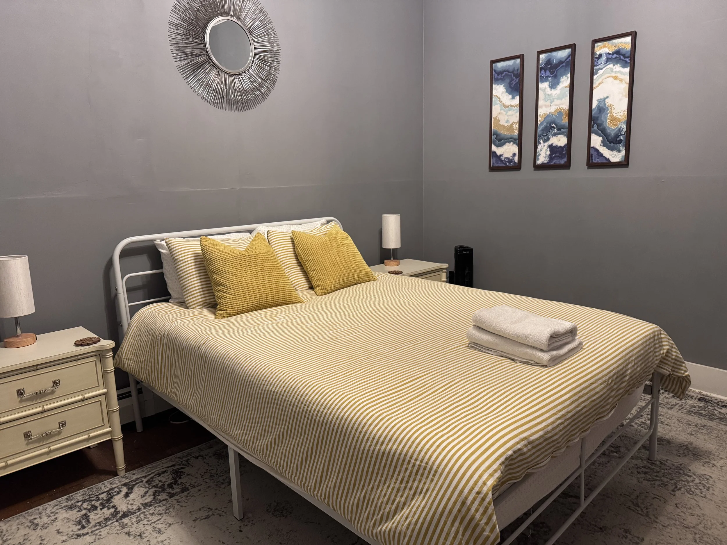A bedroom with a bed covered in yellow and white striped bedding, three matching yellow throw pillows, and folded towels on the foot of the bed. There are two cream-colored nightstands with lamps, a mirror with a sunburst frame on the wall, and three