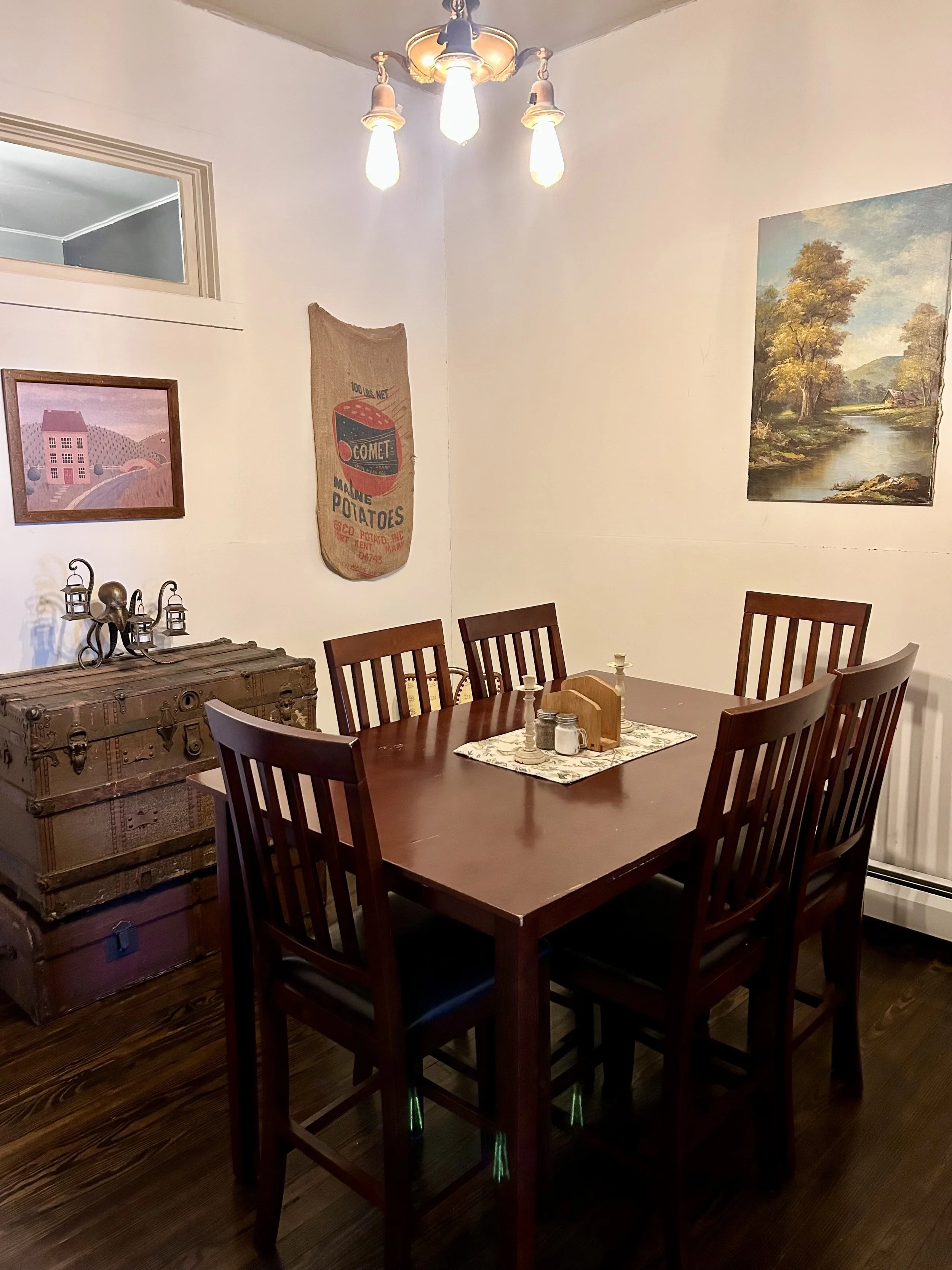 A dining room with a wooden table and six matching chairs, a decorative antique trunk with lanterns and a sculpture on top, artwork on the walls including a landscape painting and a vintage potato sack, and a chandelier with exposed bulbs hanging fro