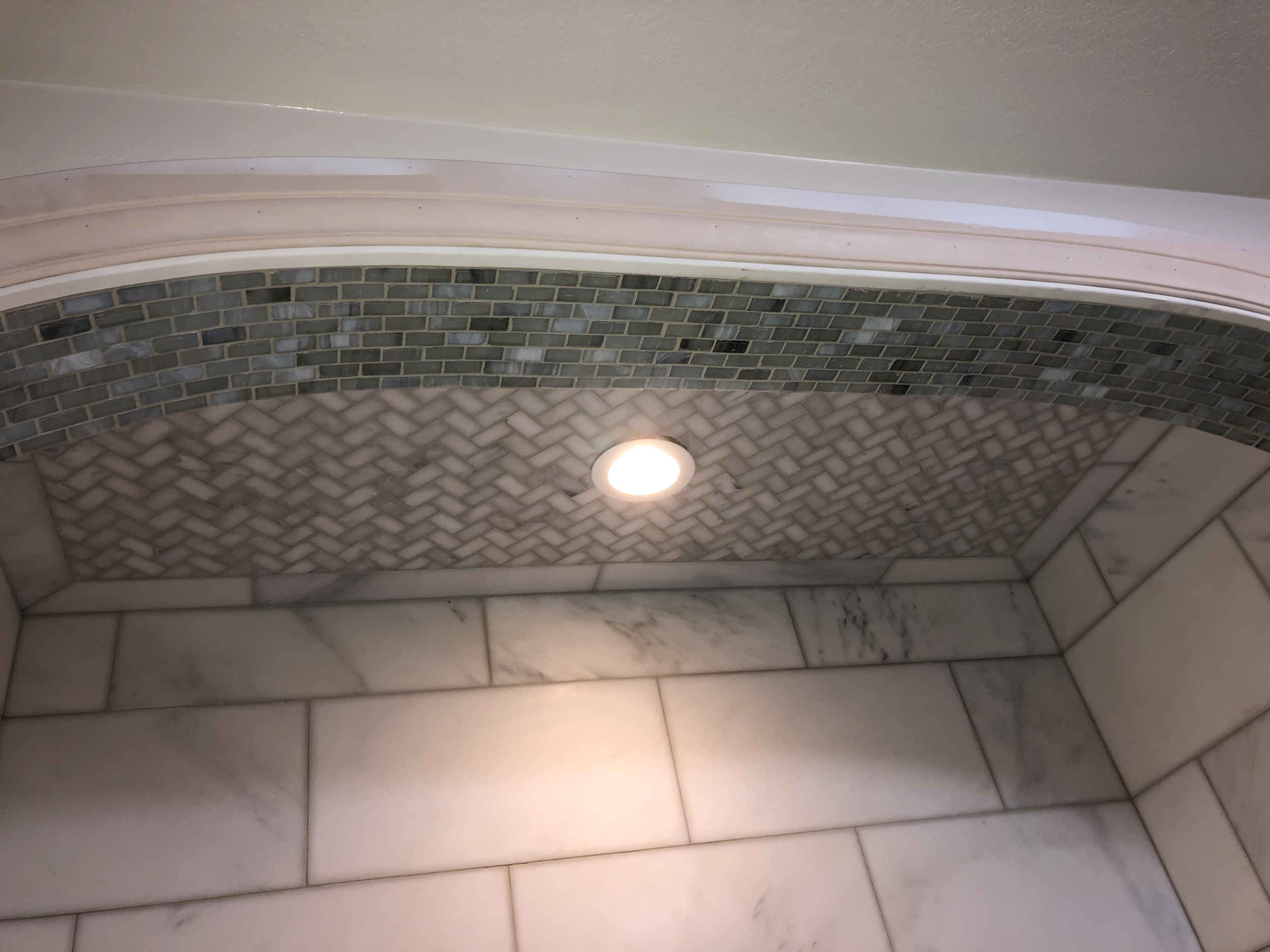 Close-up of a bathroom shower area with a curved wall with small gray and black mosaic tiles and larger white marble tiles on the lower part, illuminated by a recessed ceiling light.