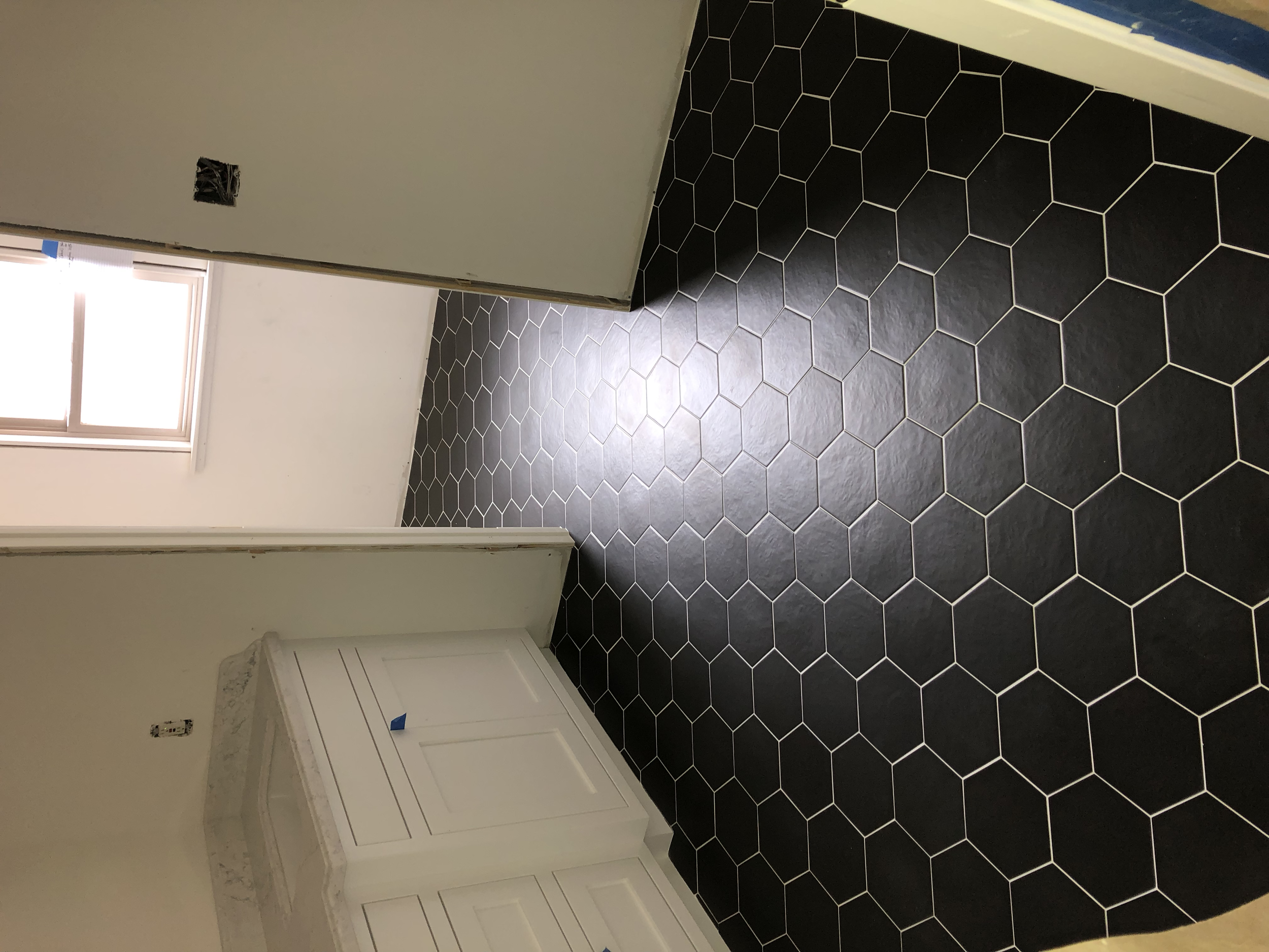Room with white walls, black hexagonal floor tiles, a window letting in natural light, an unfinished wall outlet, and a white cabinet with marble top.