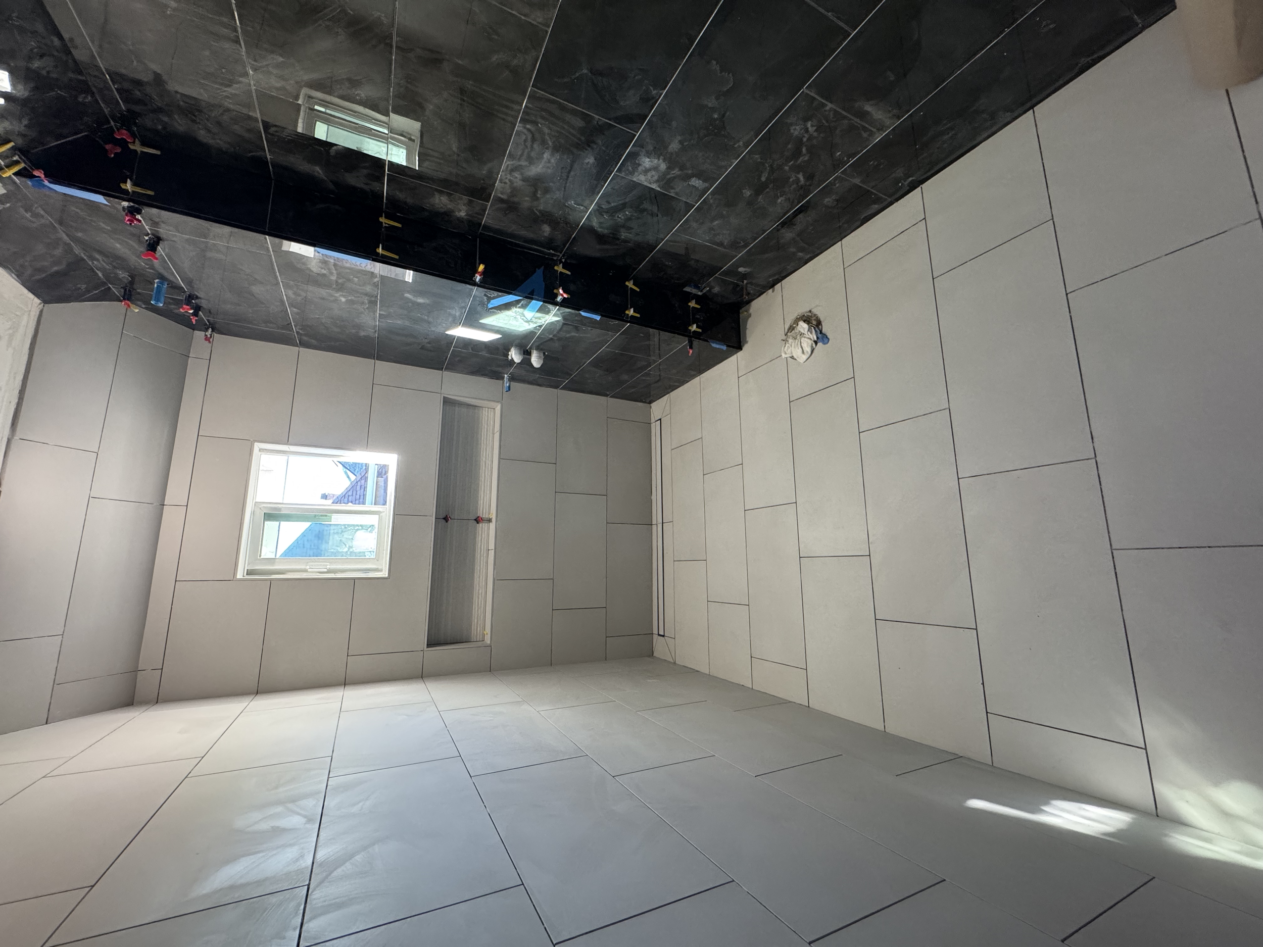 Room with white tiled walls and floor, black ceiling, a small window, and construction materials or tools on the floor and ceiling.