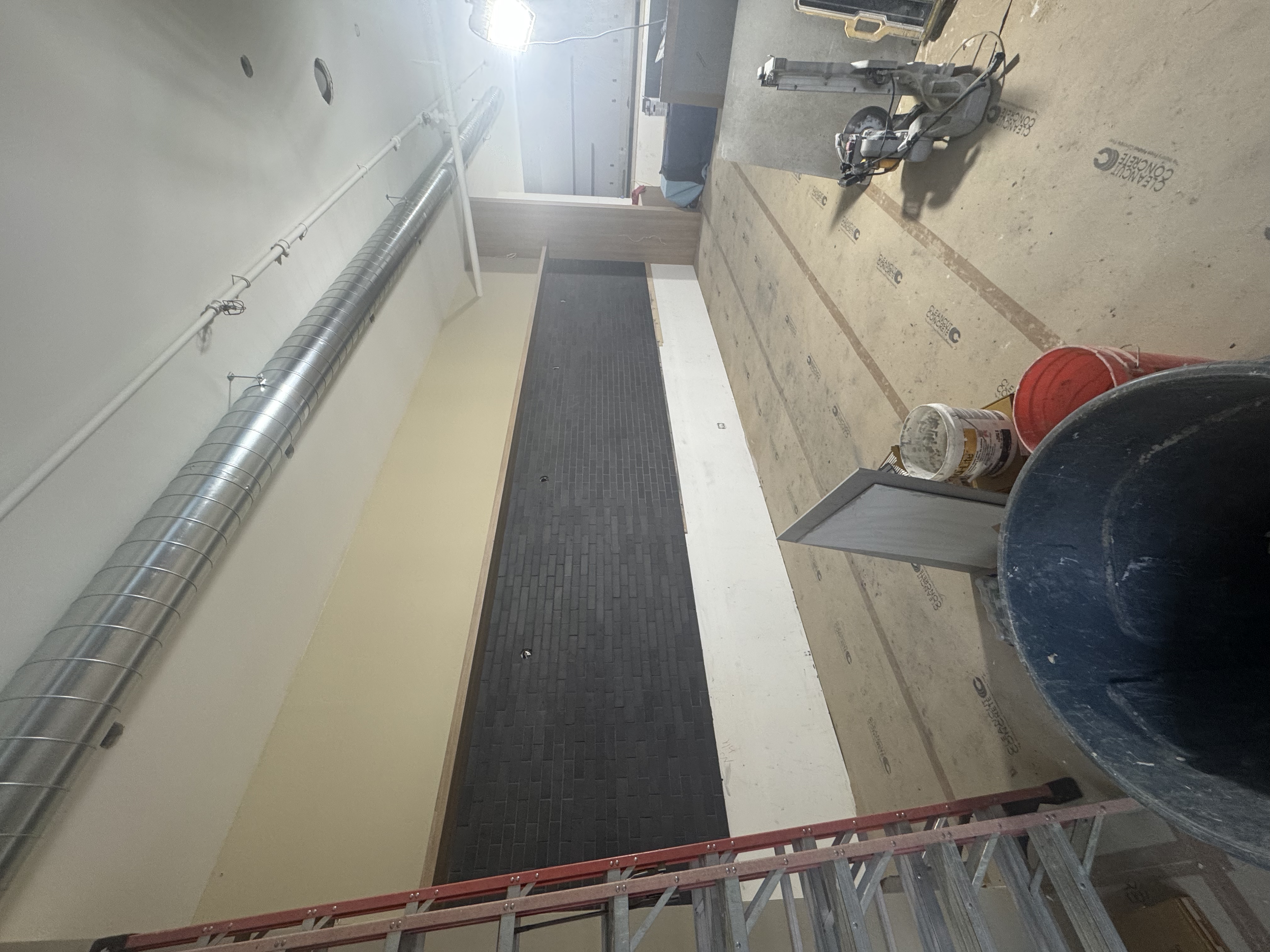 Construction site with a partially finished wall, a metal ladder, construction tools, a bucket, and a large round pipe in an interior space under renovation.