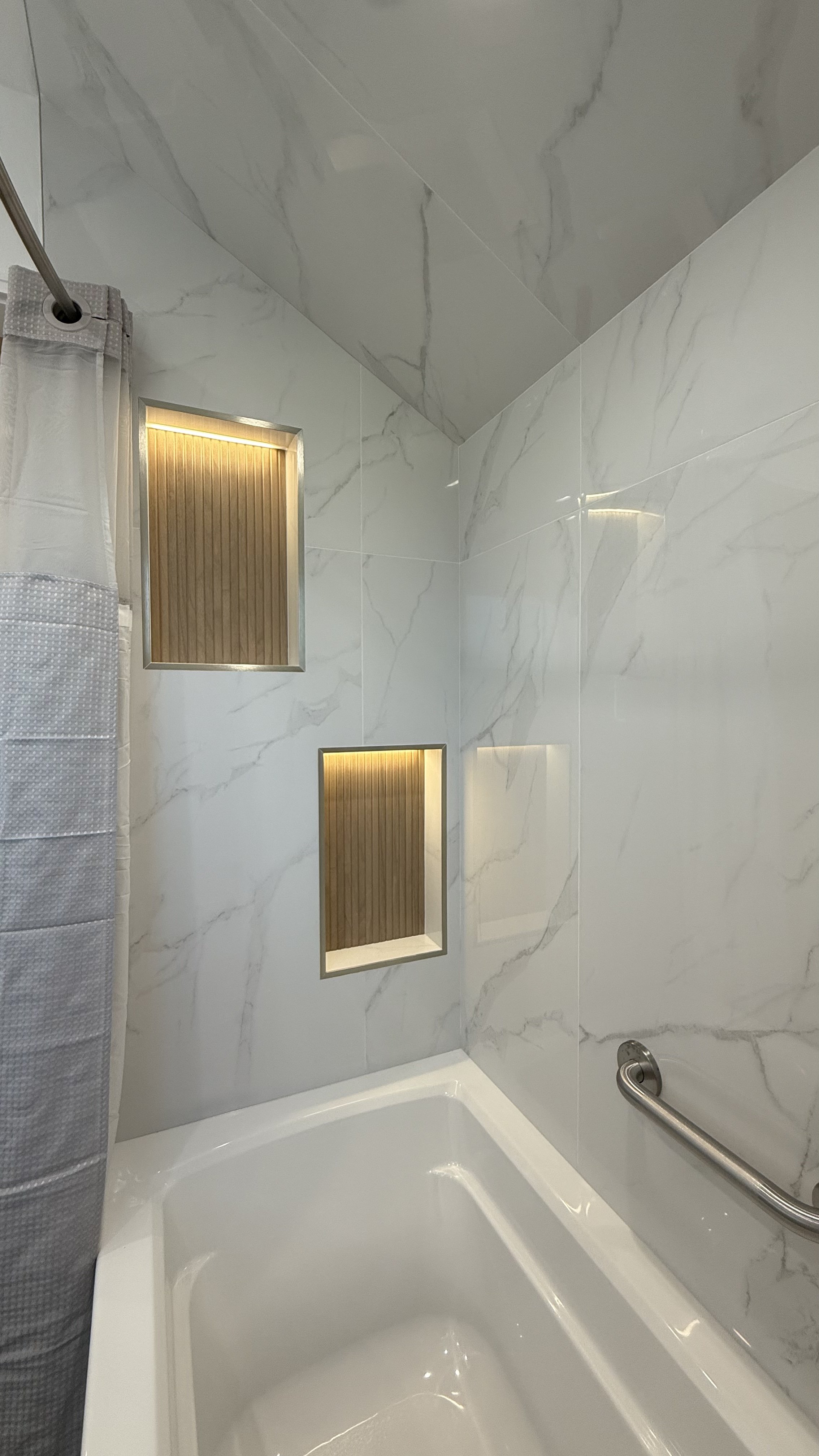 Close-up of a modern shower with white marble tile walls, built-in recessed shelves with wood backing and LED lighting, a silver grab bar, and a shower curtain on the left side.