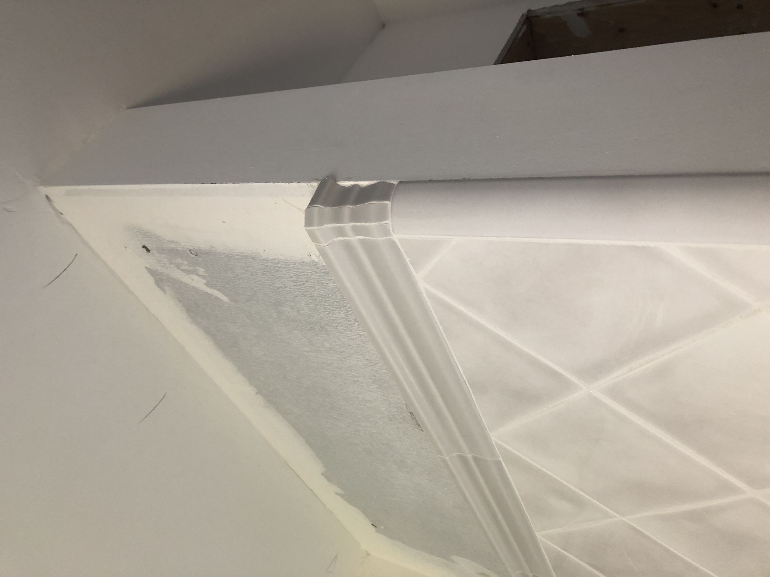 Interior view of a ceiling corner where drywall meets tiled wall, with visible tape and unfinished areas.