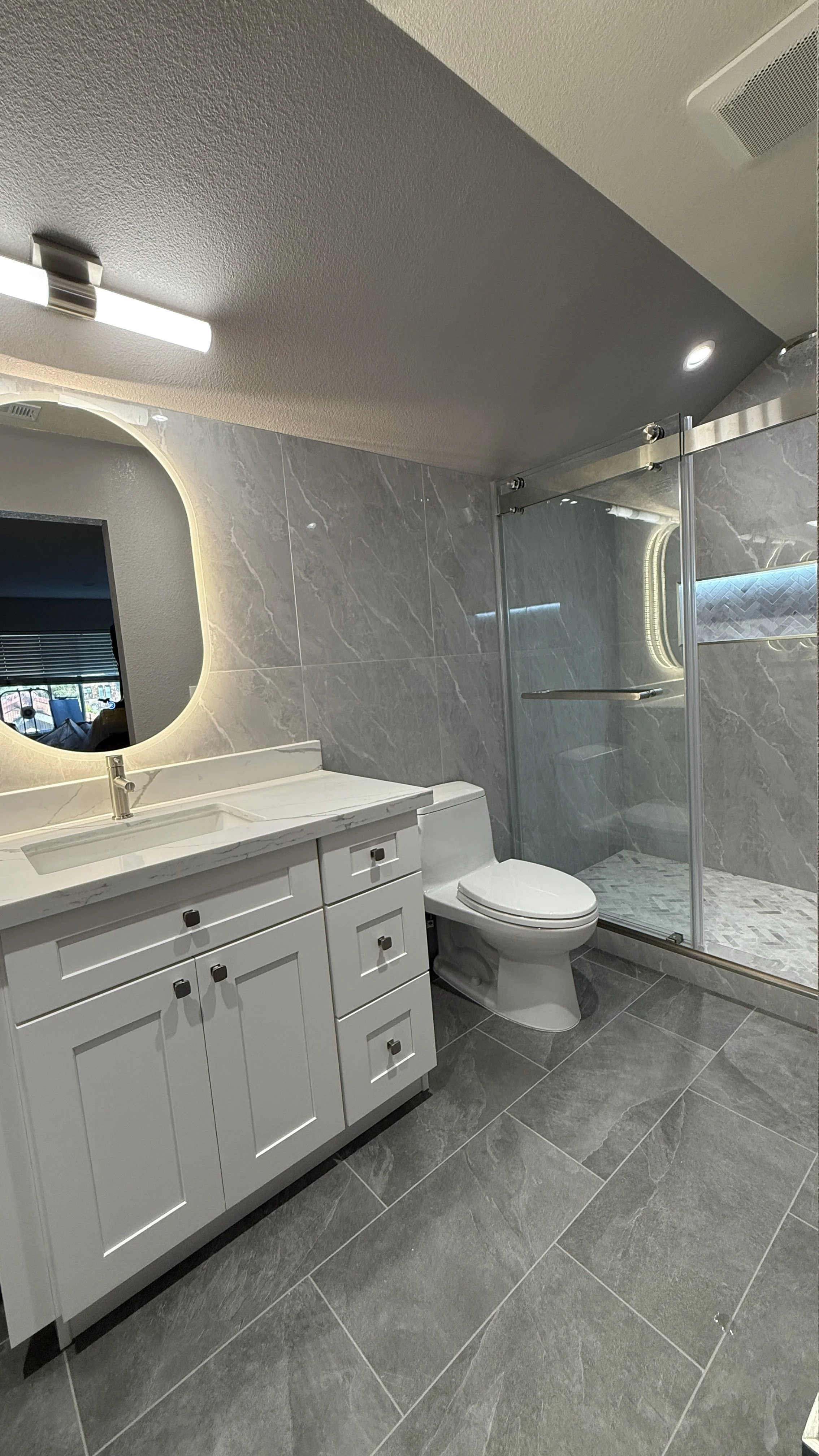 A modern bathroom with a gray and white color scheme. It features a white vanity with drawers, a marble countertop, a rectangular sink, a mirror with backlighting, a toilet, and a walk-in shower with glass doors and gray marble walls and flooring.