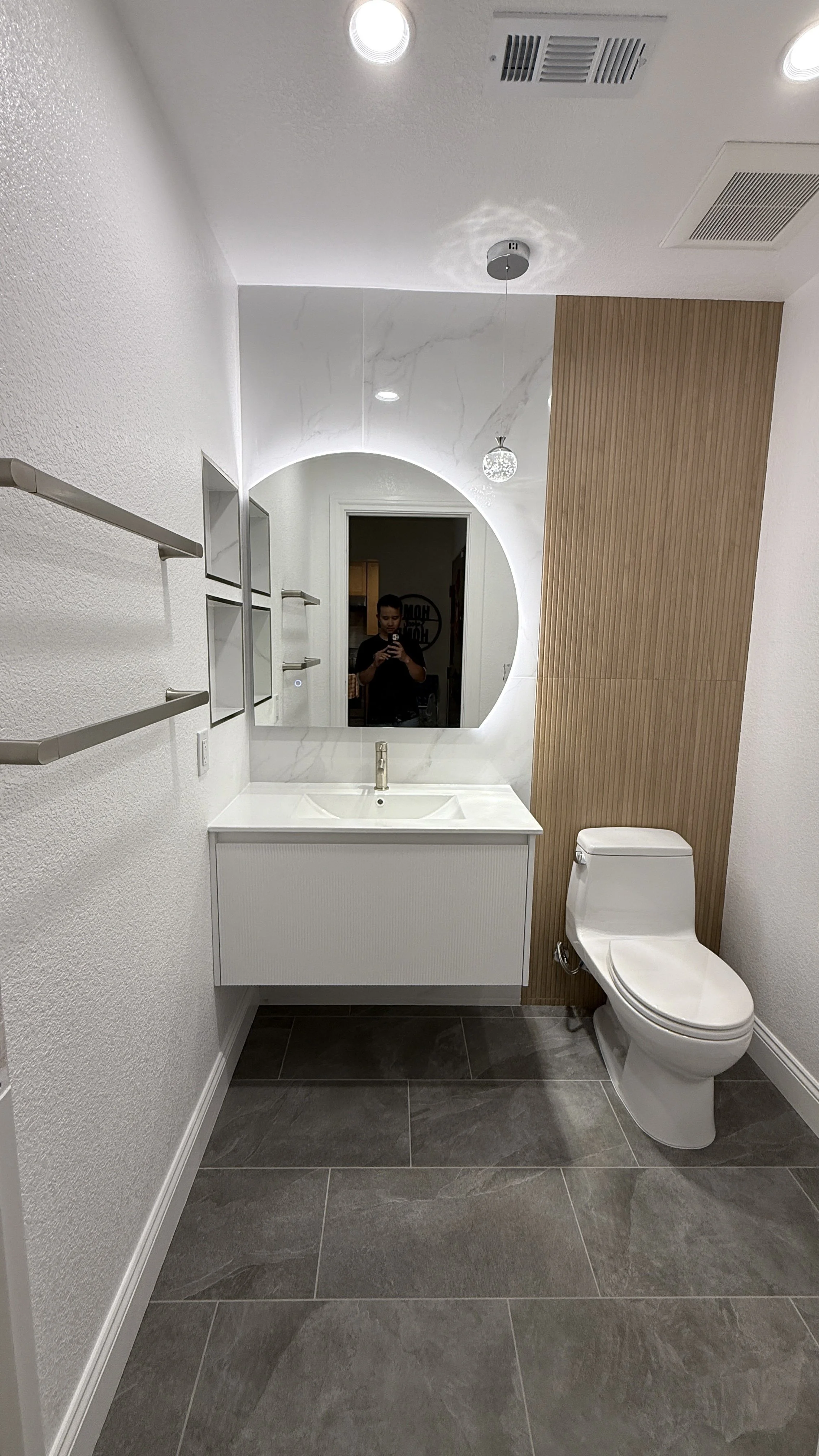 Modern bathroom with white sink vanity, large round mirror, and a white toilet. The wall behind the toilet features vertical wood paneling, and there are recessed ceiling lights and a small hanging light fixture. The floor is covered in large gray ti