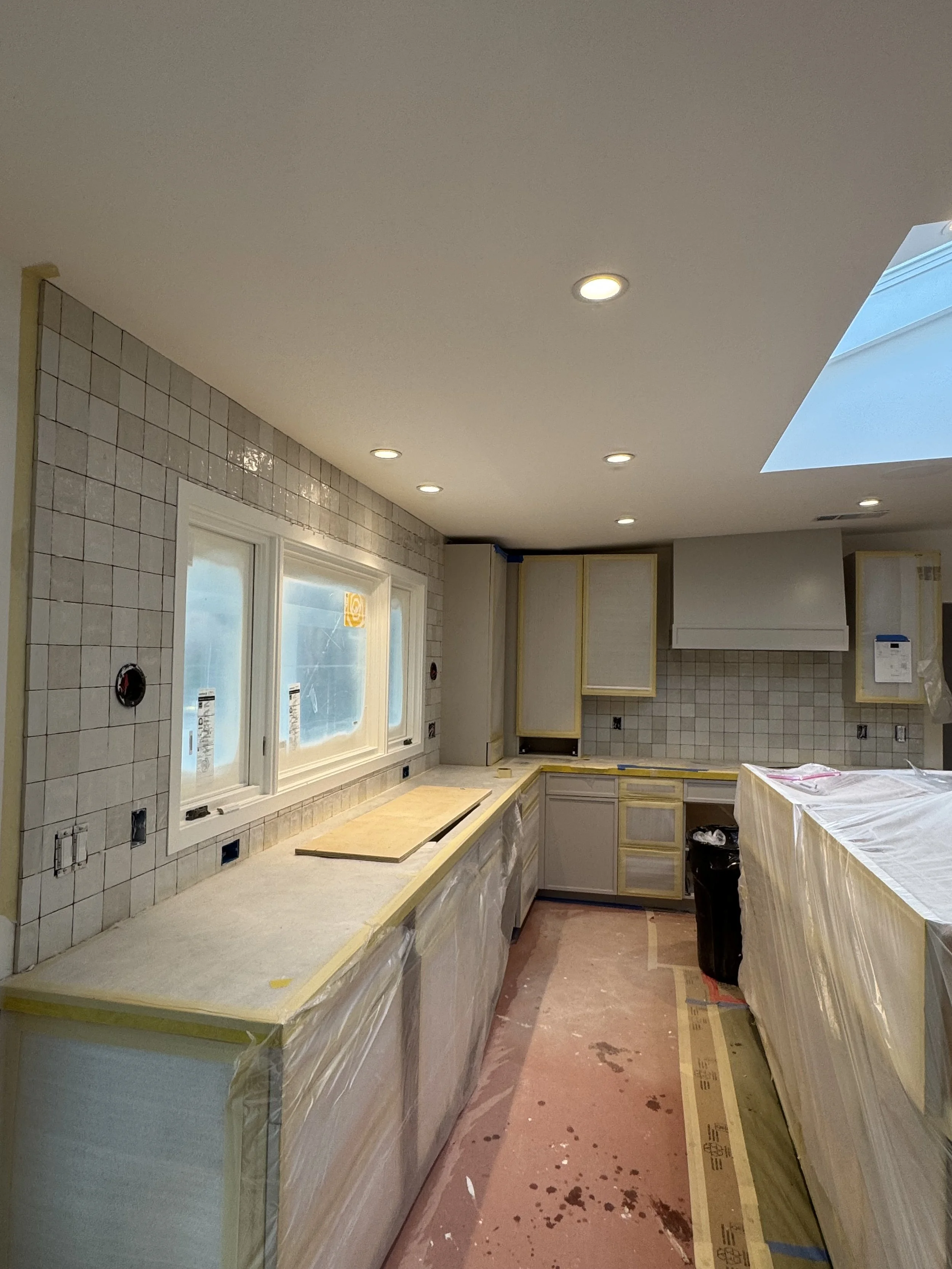 Kitchen under renovation with beige cabinets, tiled backsplash, covered counters, and a window over the sink. Protective plastic covers on cabinets and countertops, construction marks on the floor.