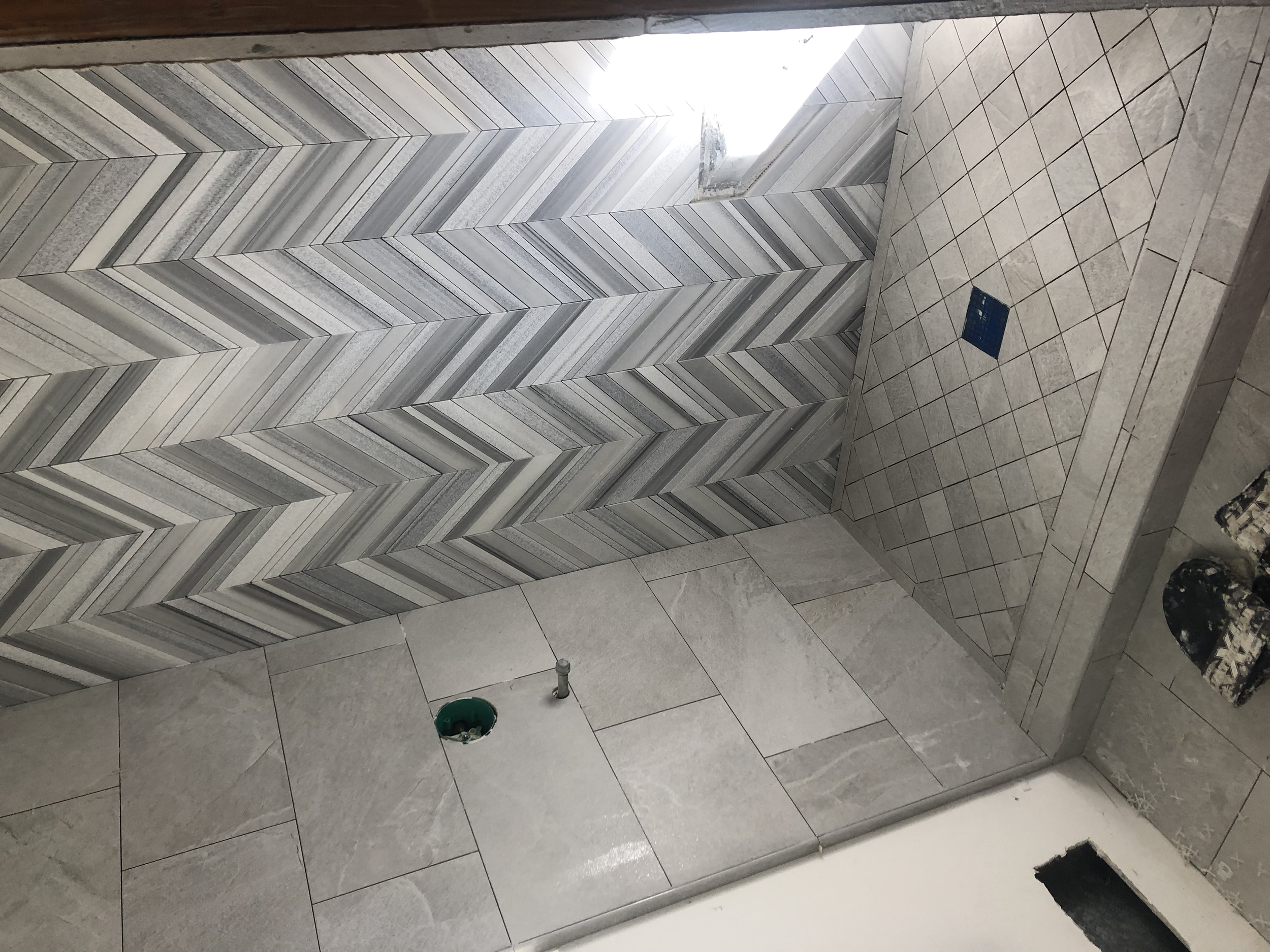 Bathroom in construction with tiled walls, including a herringbone pattern on one wall, and a floor with grey tiles. An access hole and plumbing fixtures are visible on the floor.