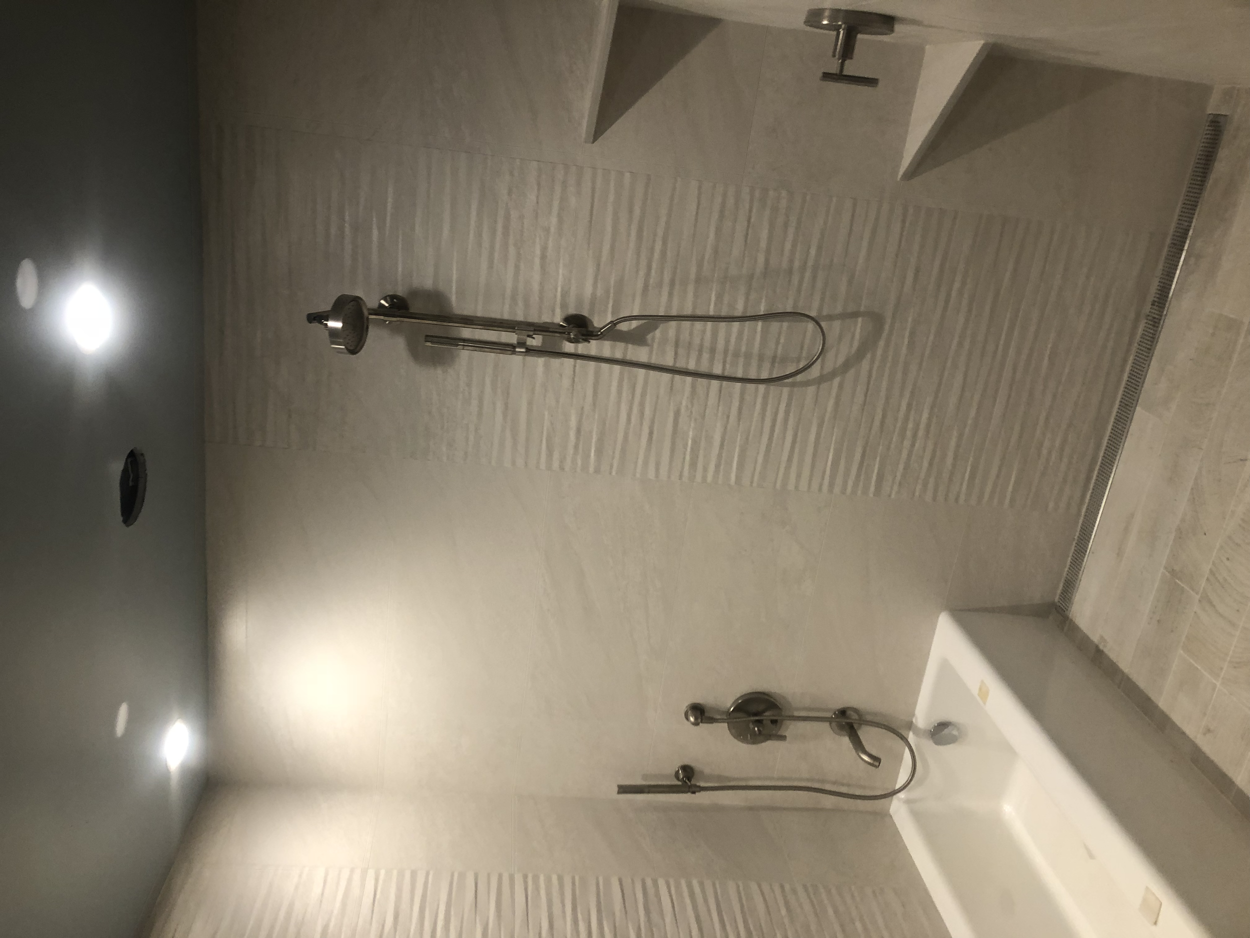 A modern shower area with a wall-mounted showerhead, a handheld shower attachment, and a small bathtub, all in a neutral beige and grey color palette, with recessed ceiling lights.