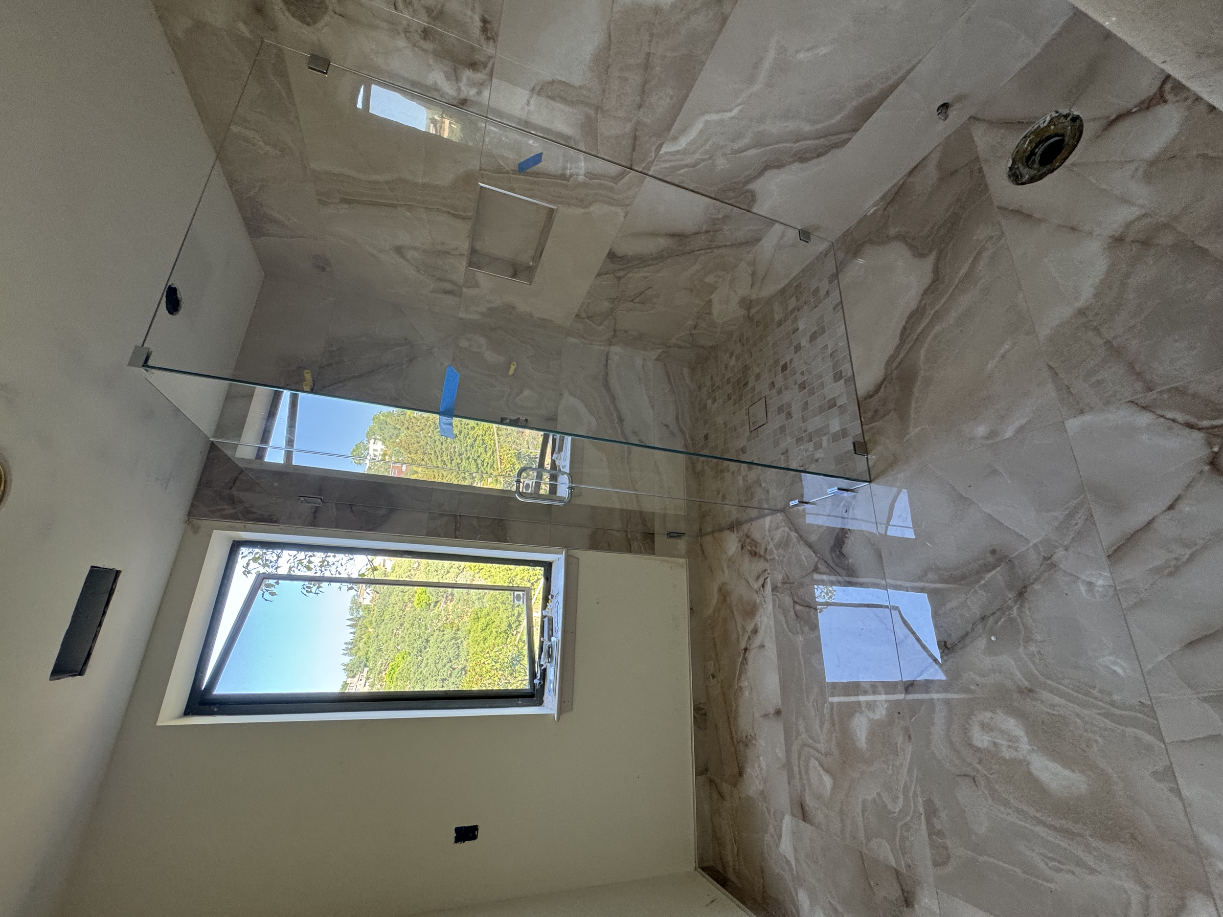 Unfinished bathroom with a glass shower enclosure, large beige marble tiles on the walls and floor, an open window showing trees outside, and no fixtures installed yet.