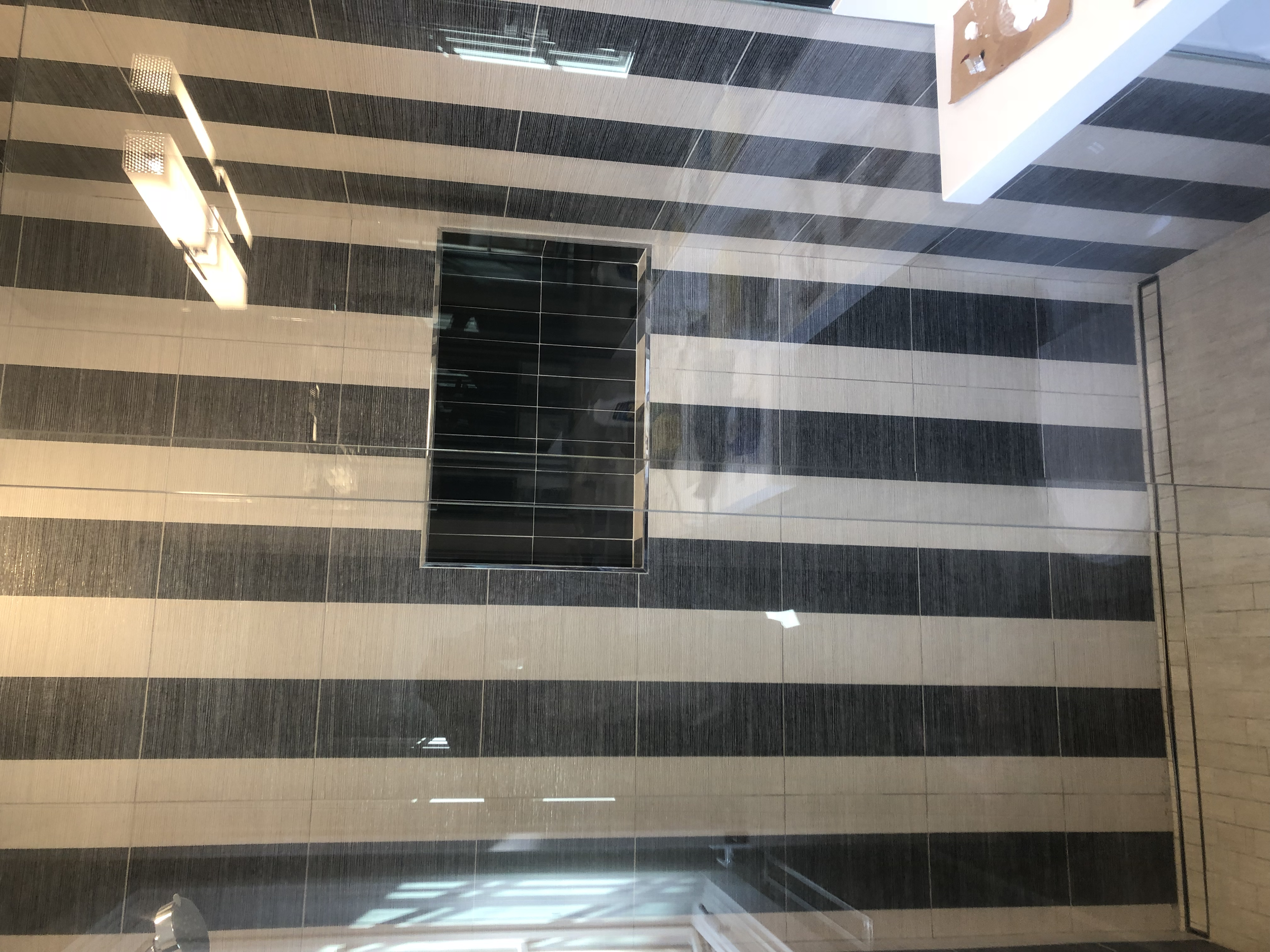 Interior view of a modern building with a striped black and white tiled wall, a large window with a brown frame, and a ceiling with hanging lights.