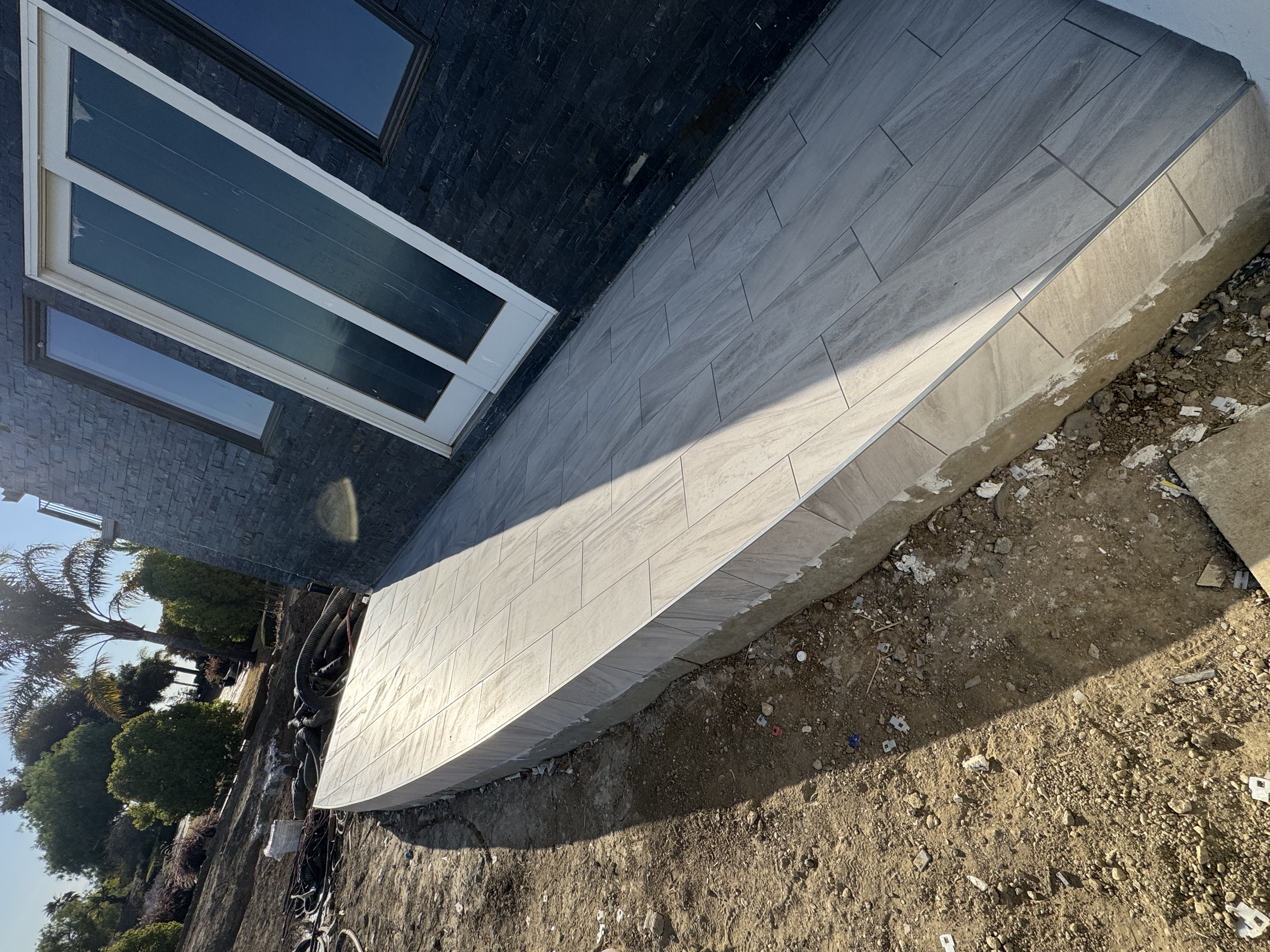 Exterior of a building under construction with a wall of black bricks, a large glass door, and a curved tiled porch area. Dirt and construction debris are in the foreground.