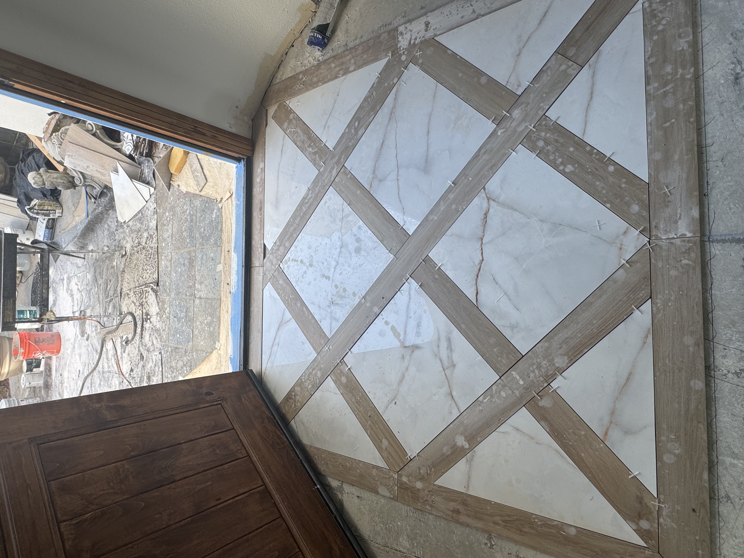 Interior view of a tiled floor under construction with a wooden frame and diagonal tiles. Outside a door, construction tools and materials are visible.