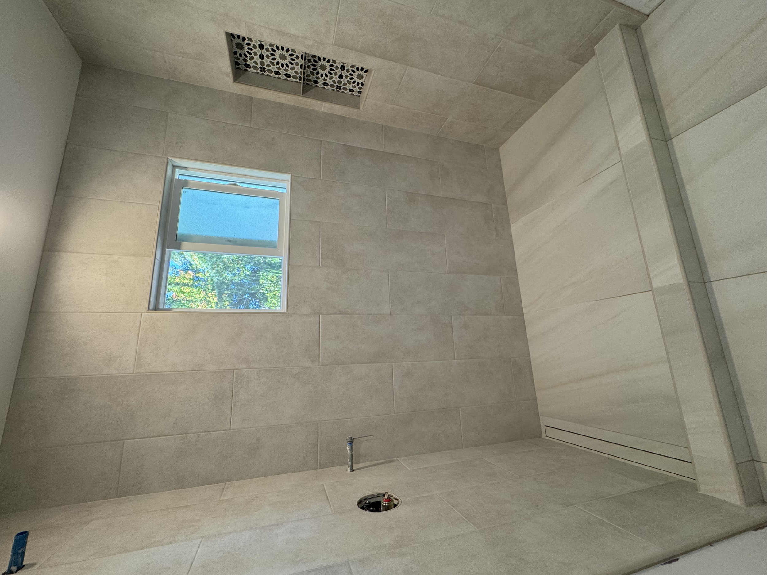  bathroom with beige tile walls and floor, a small window with a view of green trees, and a ceiling vent with decorative black and white pattern.