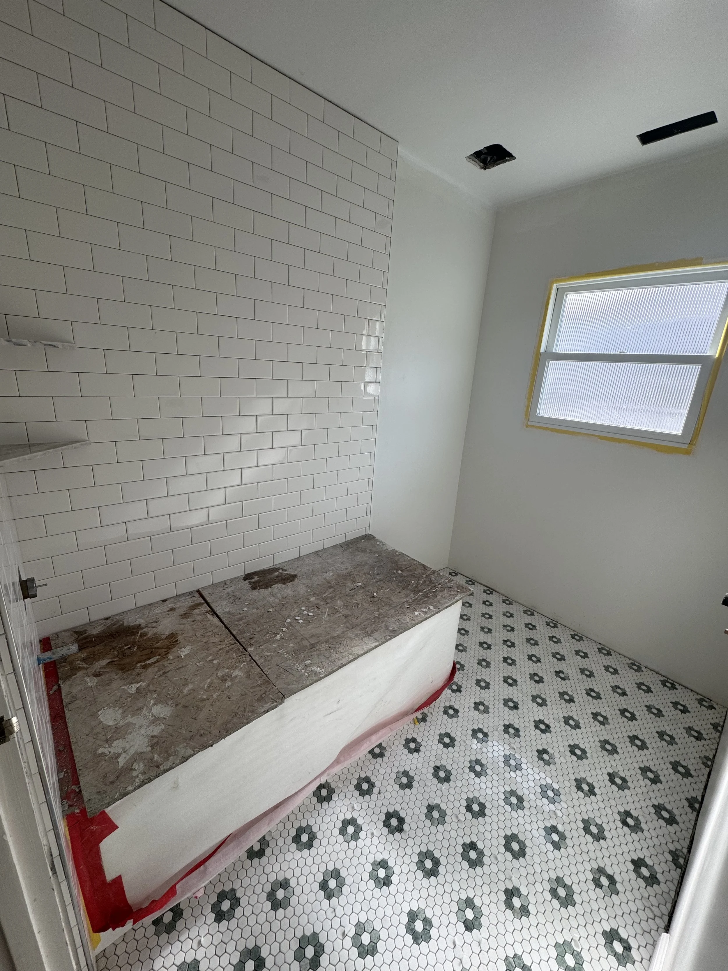 Bathroom under construction with a white subway-tile wall, a window with yellow tape, a partially covered bathtub or countertop, and black and white patterned tile flooring.