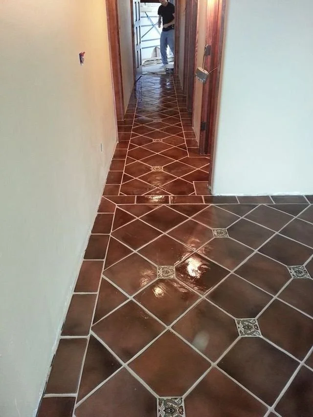 A hallway with shiny, brown ceramic tile flooring featuring decorative square accents, white walls on the left and right, and a partially open door at the end revealing a person standing outside.