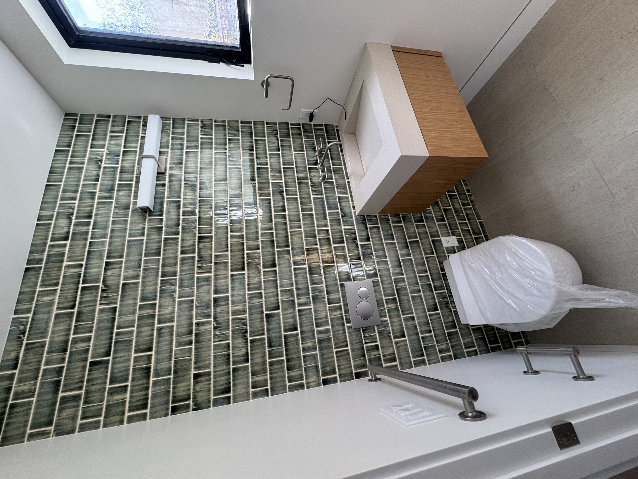 Interior of a modern bathroom with green tile wall, a window, a wall-mounted sink, a toilet with a plastic cover, and a wooden cabinet.