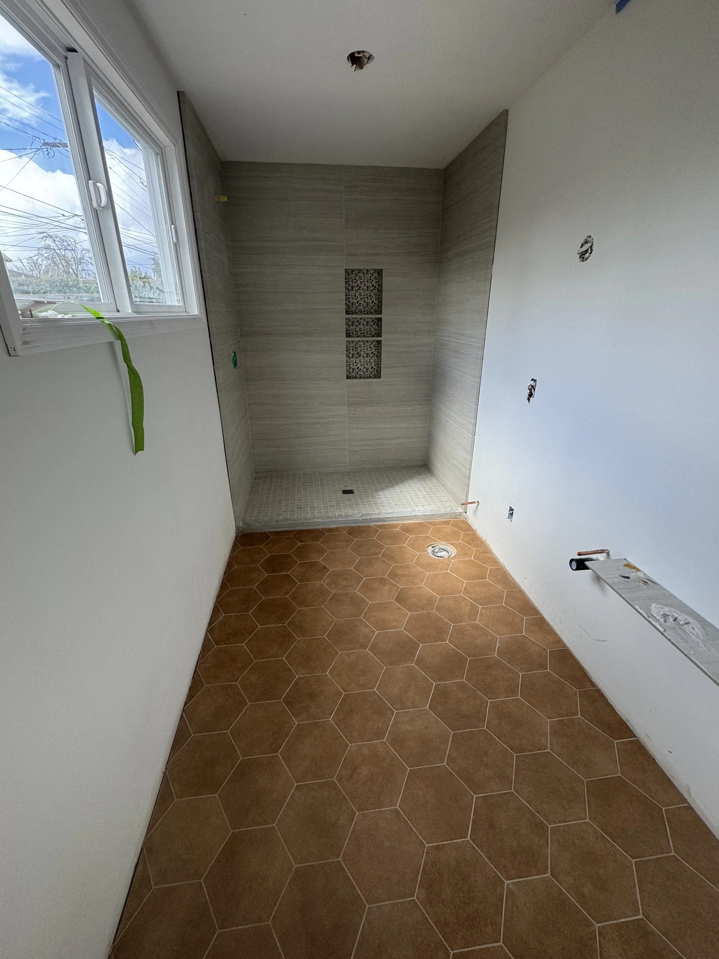Bathroom under construction with hexagonal terracotta tiles on the floor, a tile shower area with beige tiles and built-in niches, a window on the left, and unfinished walls and ceiling with visible holes for fixtures.