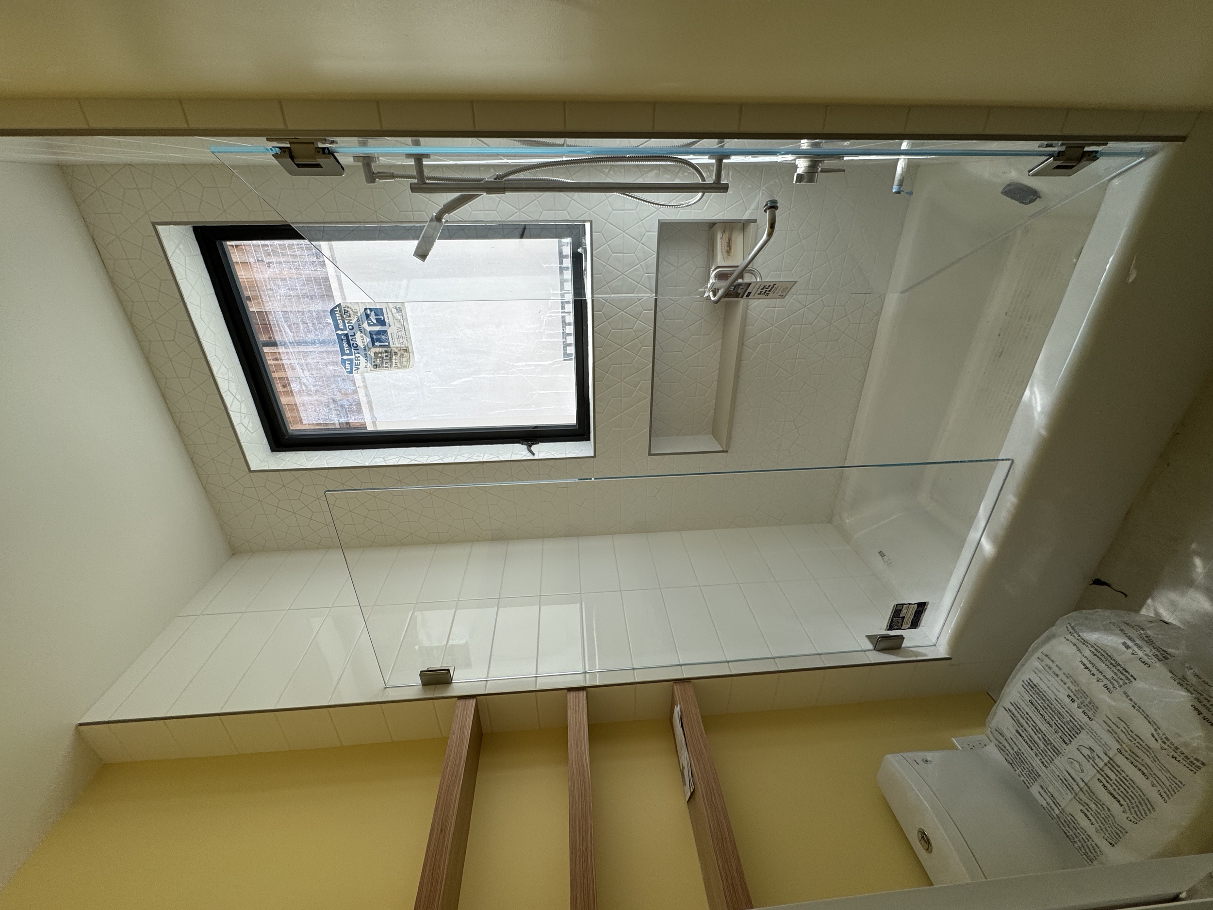 Bathroom with a glass shower enclosure, a window, and a toilet with a plastic cover, yellow walls, and wooden shelf support