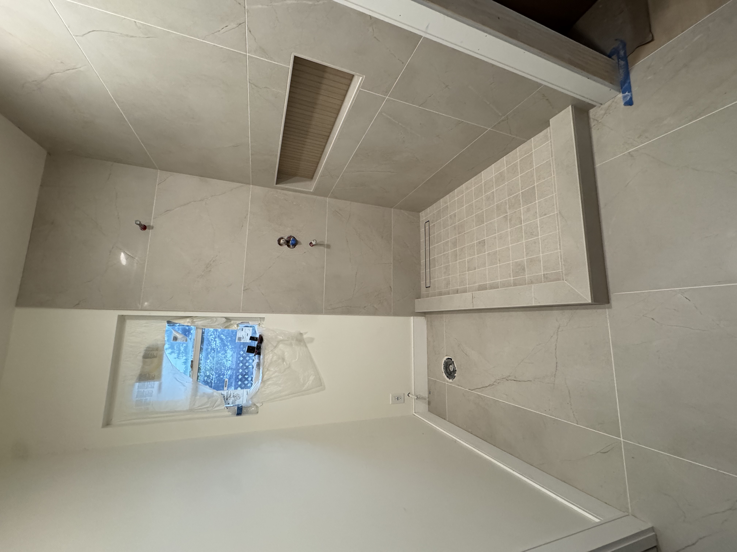 Unfinished bathroom with beige tile walls and floors, plumbing fixtures, and a recessed shower area with small beige tiles, window with plastic covering, and construction materials.