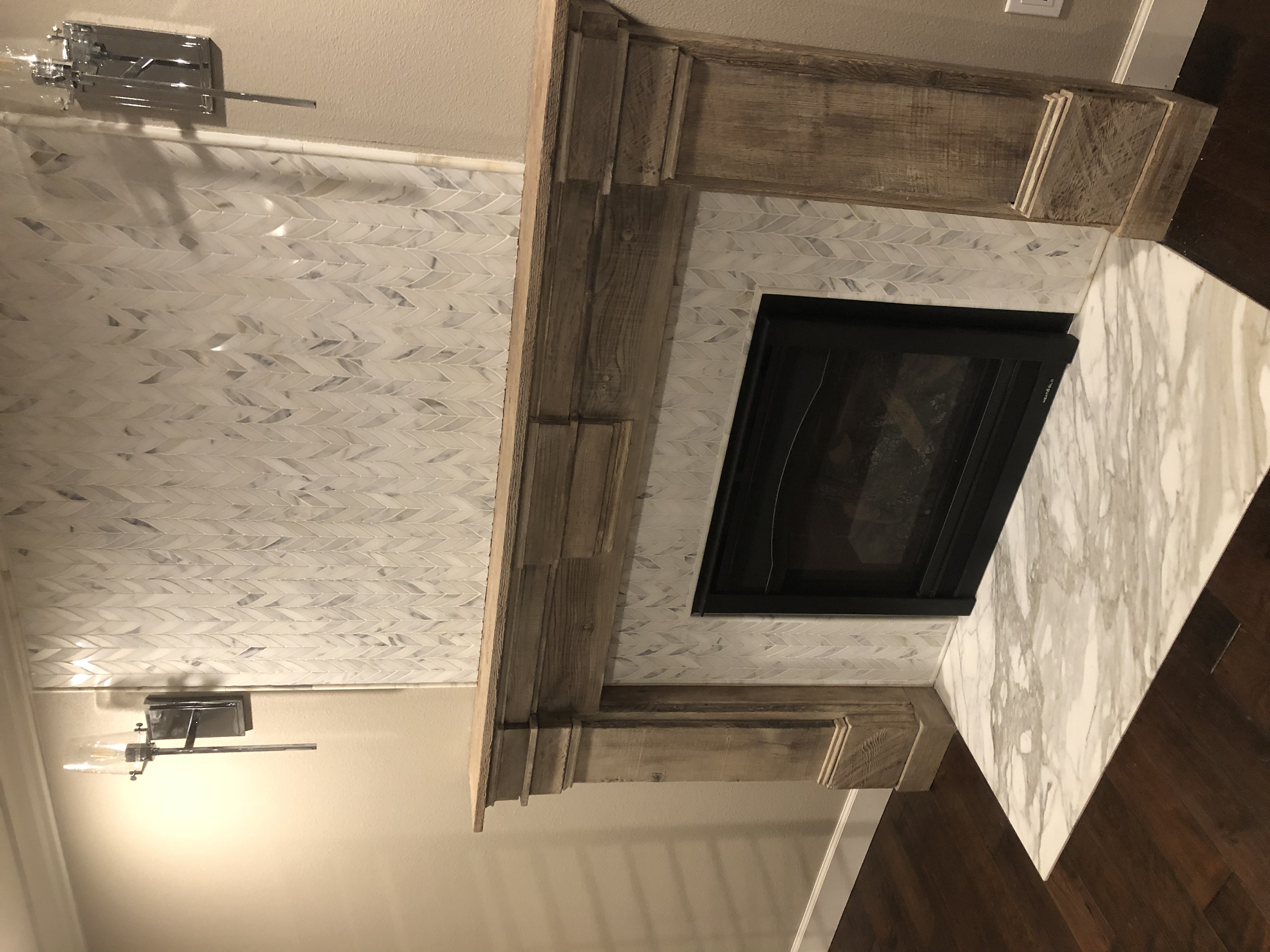 Living room fireplace with a black glass front, surrounded by a white marble hearth, with wooden cabinetry on each side and a decorative tiled wall behind. A wall-mounted light fixture is visible on the left.