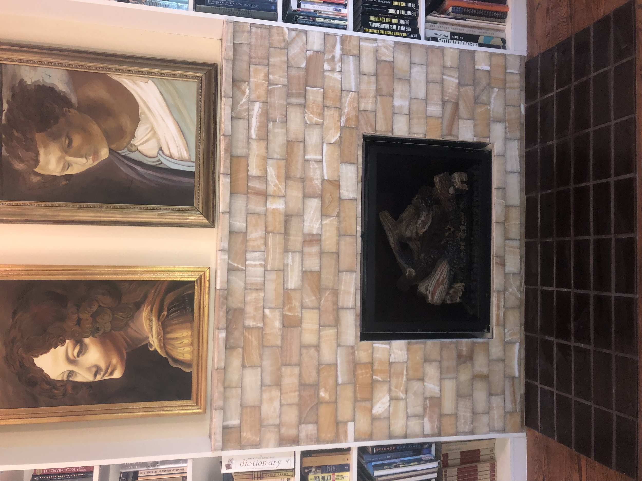 A brick fireplace with a metal fire grate and stacked logs, flanked by two framed classical style paintings of women, and a bookshelf filled with books above the fireplace.