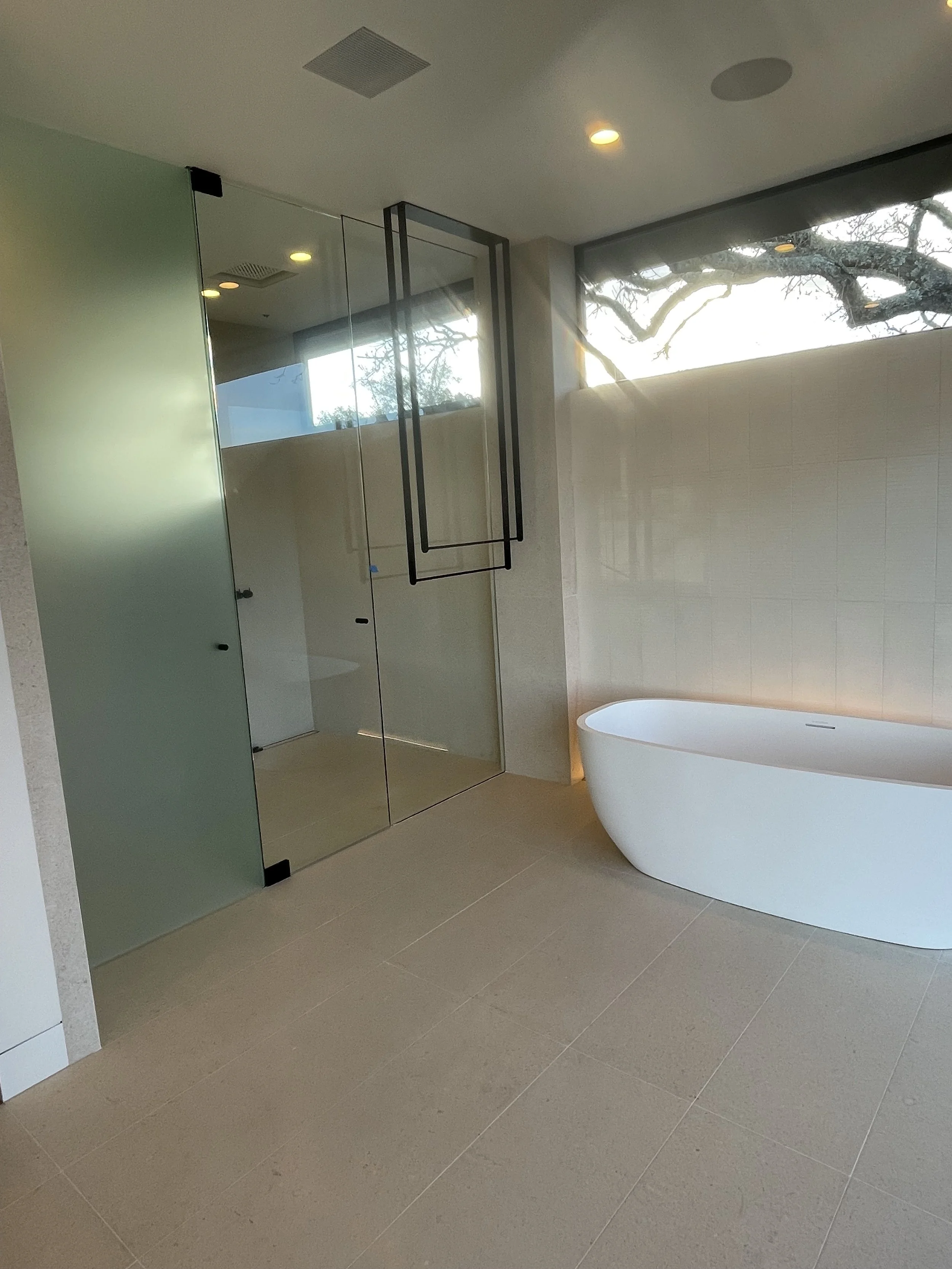 Modern bathroom with a white freestanding bathtub, tiled floor, glass shower enclosure, and a wall window showing tree branches outside.