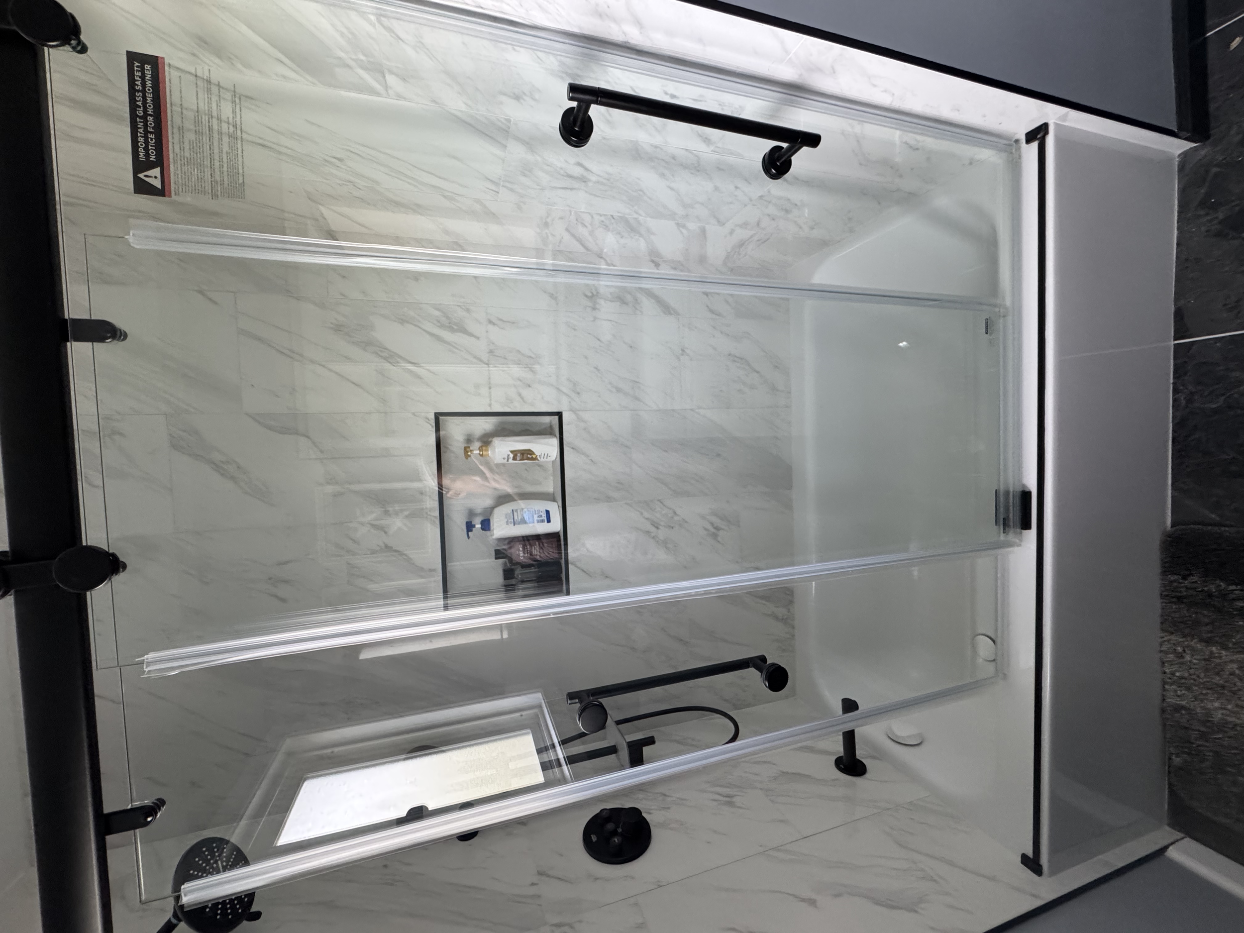 A modern glass shower enclosure with black hardware, set against a white marble wall with gray veining, in a bathroom.