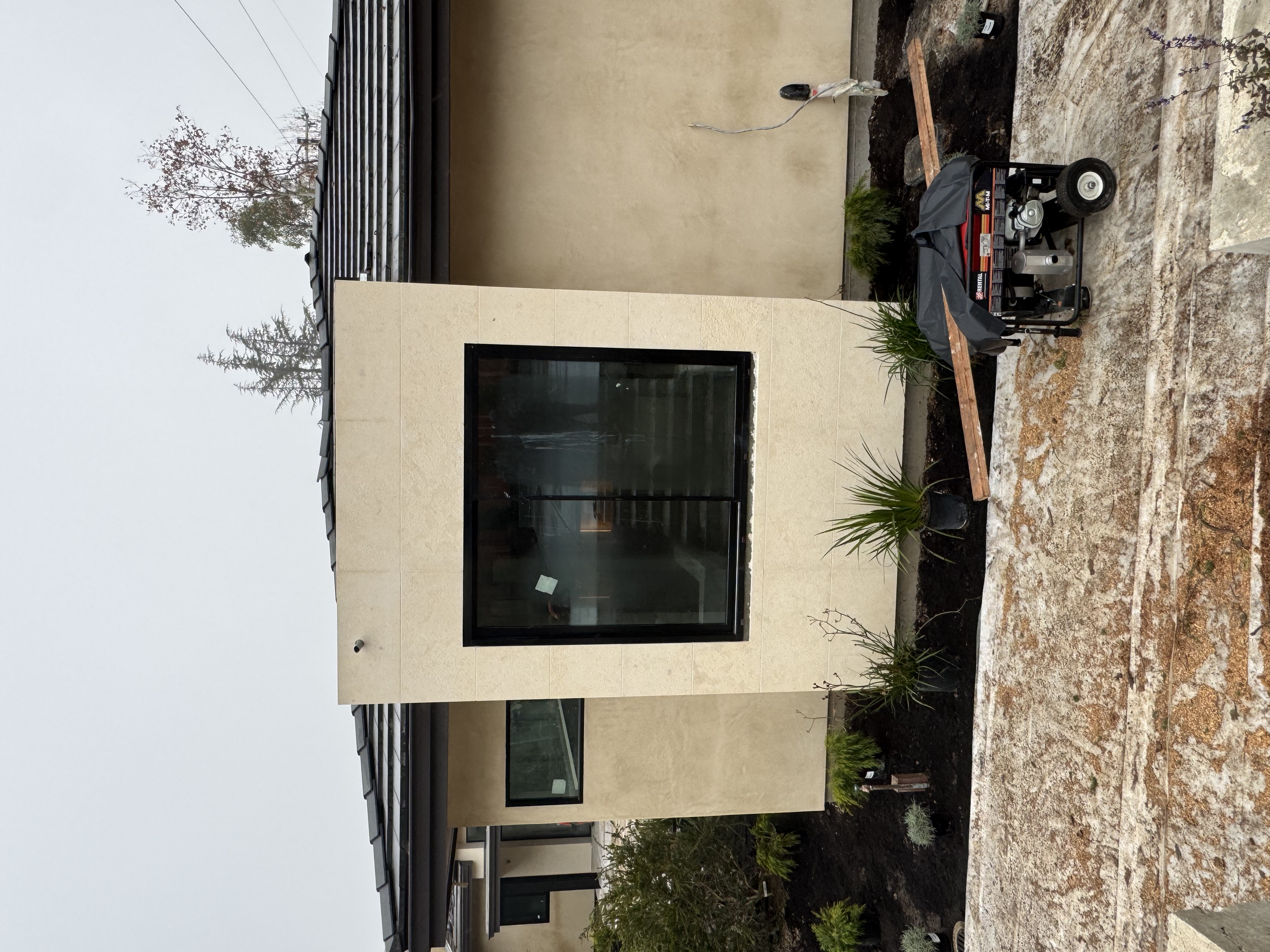Modern house exterior with a large black-framed window, small plants, and a portable generator on a raised wooden platform.