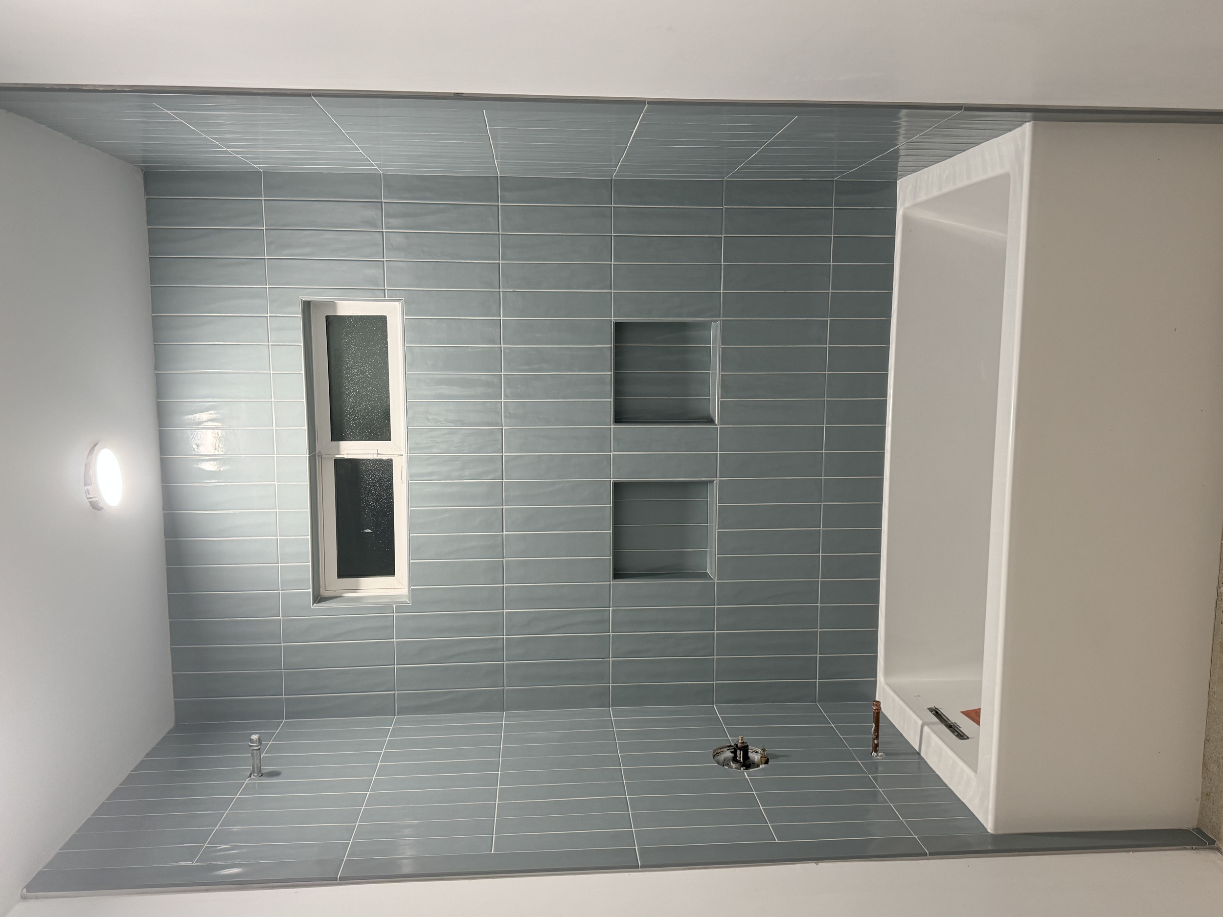 Newly constructed bathroom with blue tiled walls, a built-in bathtub, and two rectangular niches in the wall, with a frosted window above and a ceiling light.