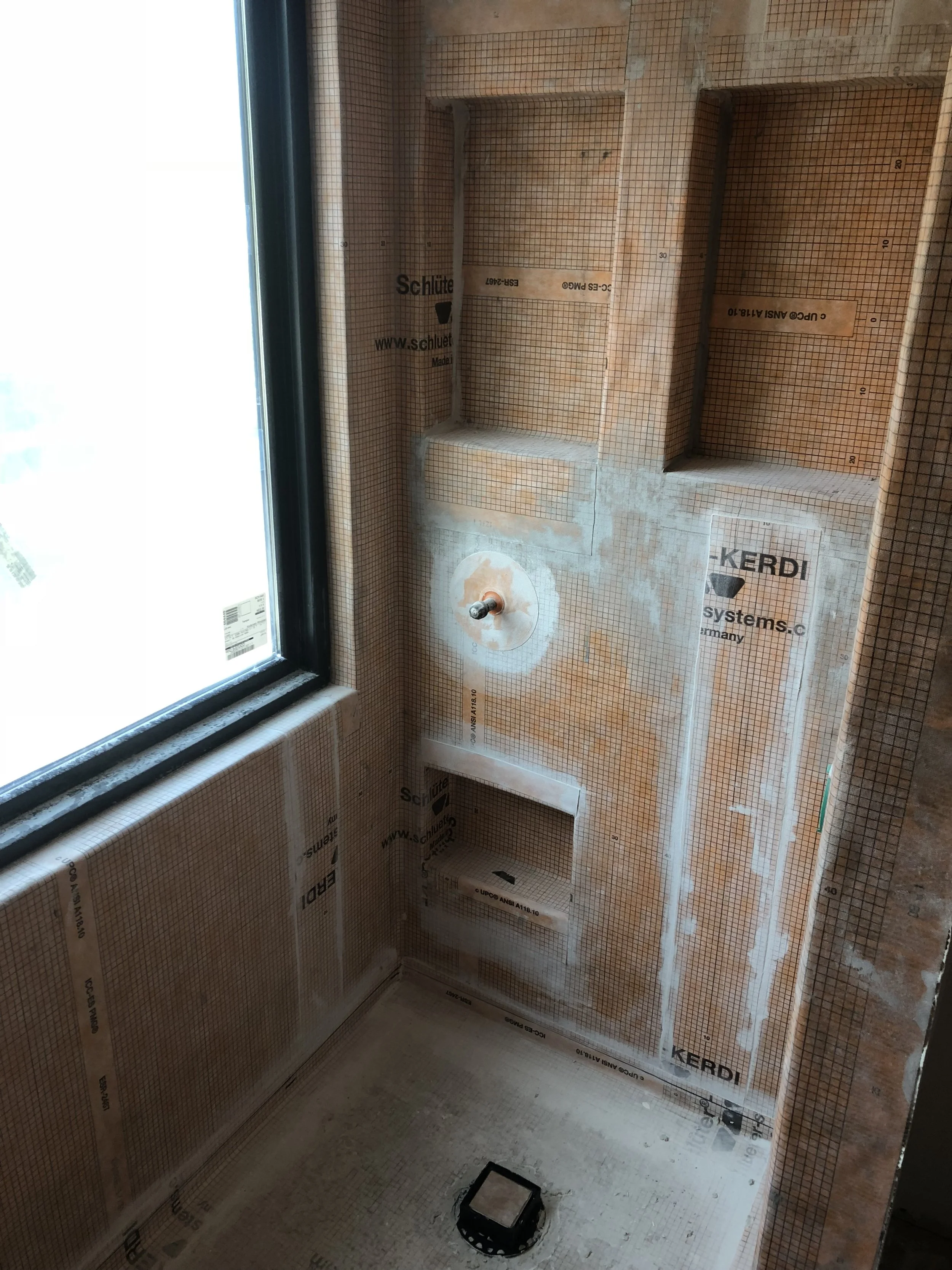 Bathroom or utility room under construction with plumbing installed, insulation and drywall unfinished. There is a window on the left and a black pipe with a flange on the wall, probably for a toilet or sink. The floor is unfinished and there is a small black square object in the middle.