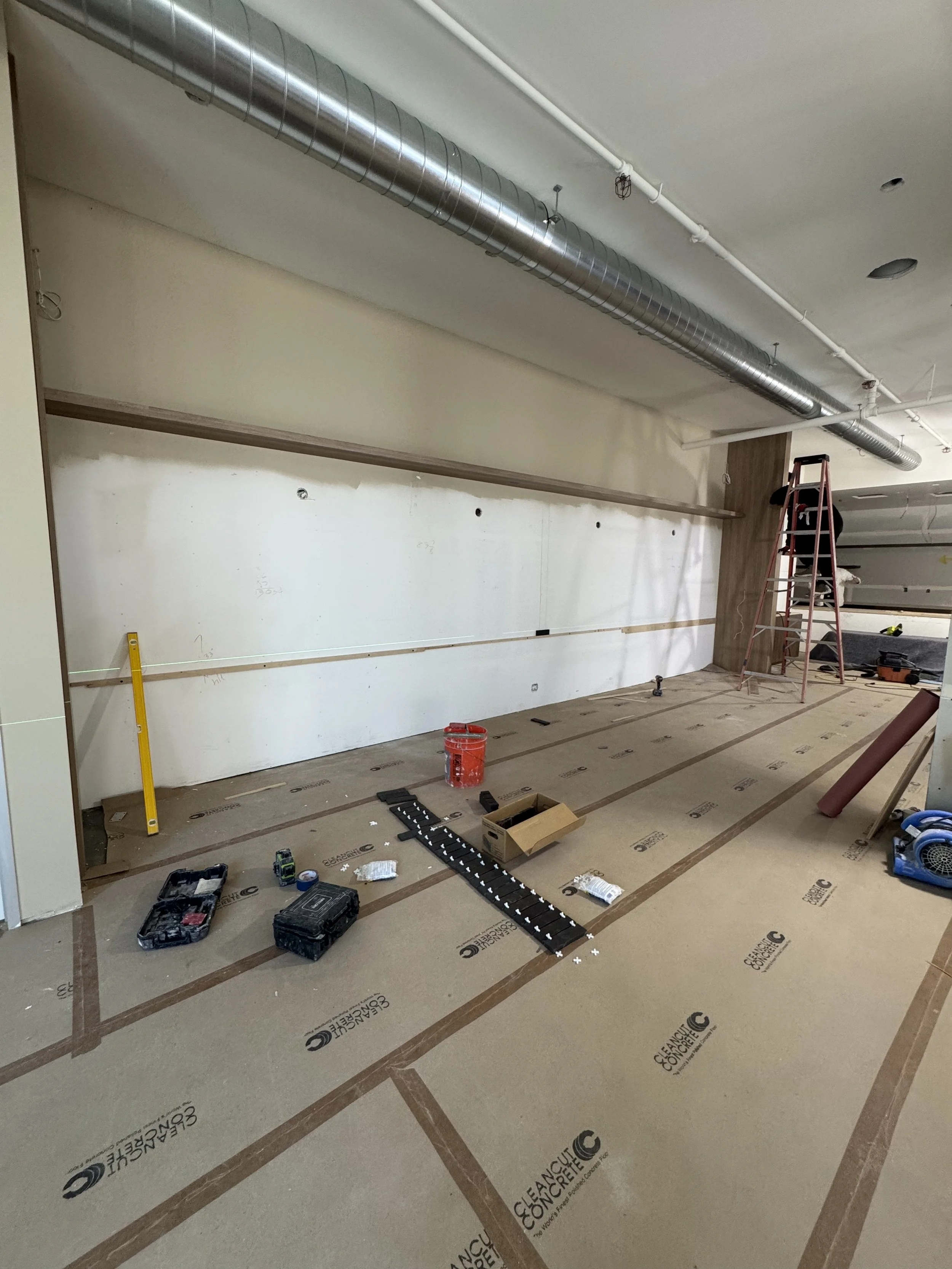 Room under construction with tools, a ladder, and protective flooring, showing ongoing wall and ceiling work.